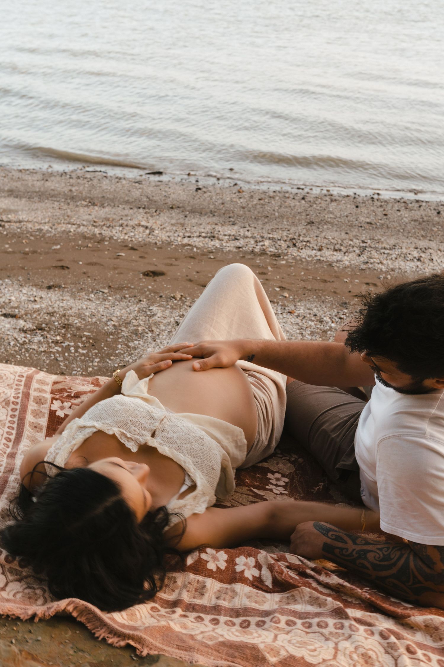 A couple lies peacefully on a patterned blanket at the beach shore during golden hour.