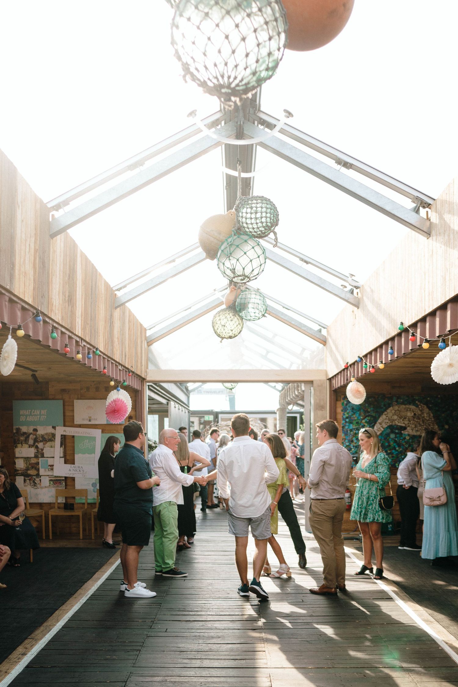People gather in a sunny indoor market hall with glass ceiling and decorative mirror ball lights hanging above the wooden walkway.