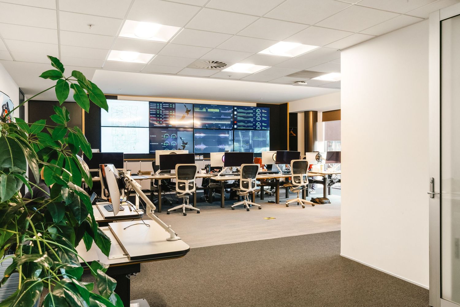 Open office workspace with multiple monitors, desks, and indoor plants in a well-lit corporate setting.