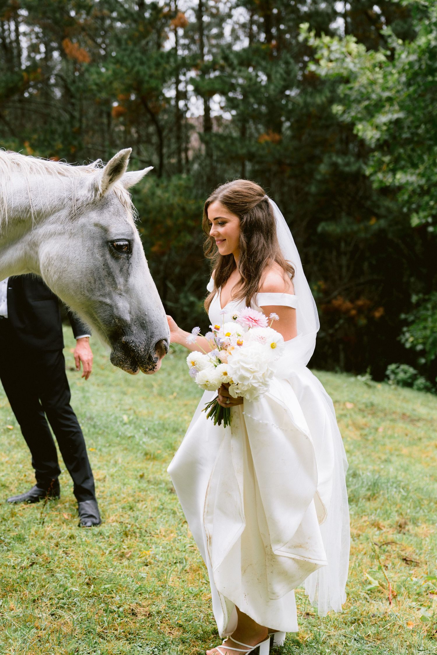 A white horse gently interacts with a bride holding a bouquet in an outdoor setting.