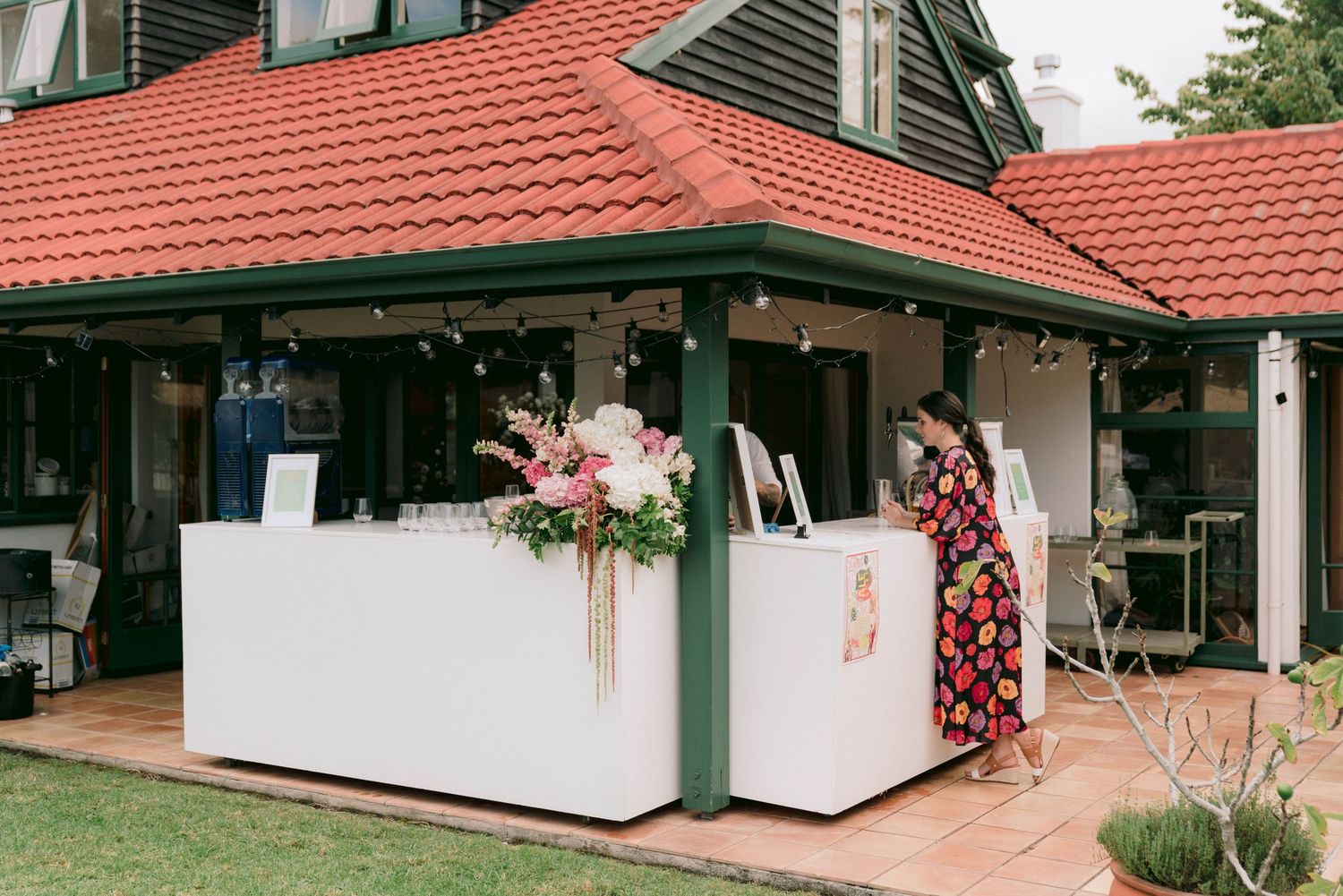 Modern white bar setup outside a house with red tile roof and floral decorations.