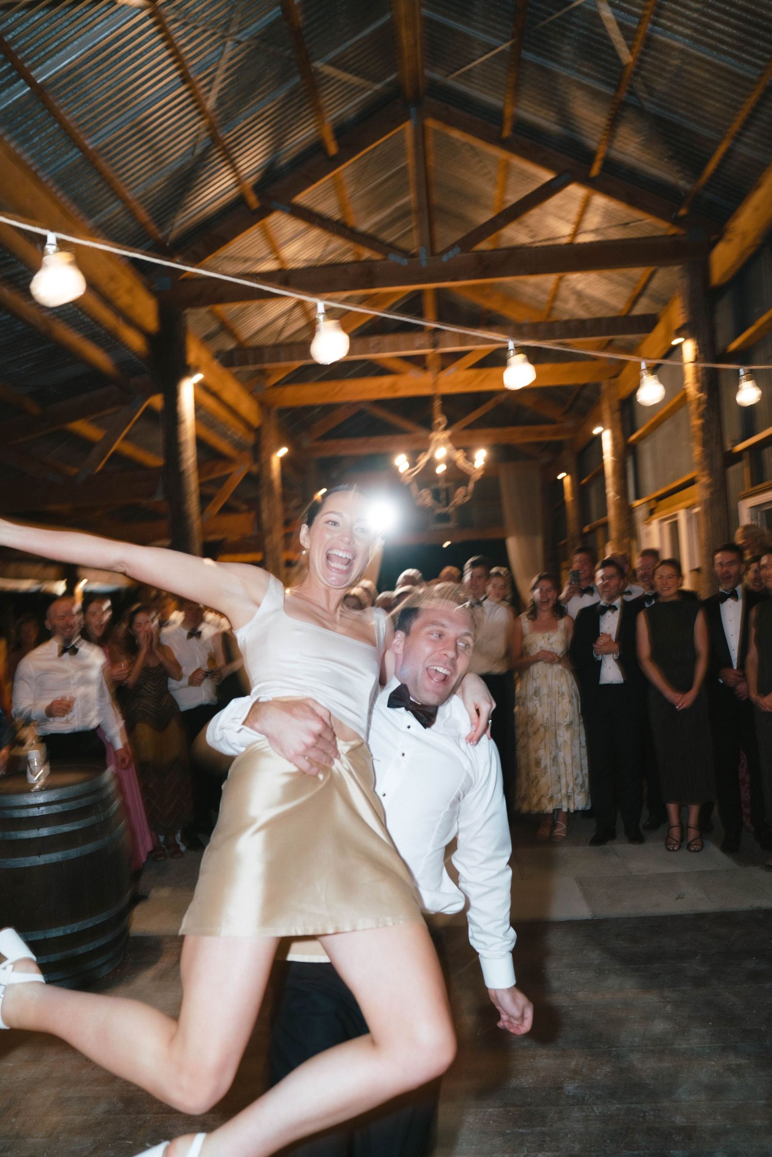 Newlyweds sharing an energetic dance moment under string lights in a rustic barn reception venue.