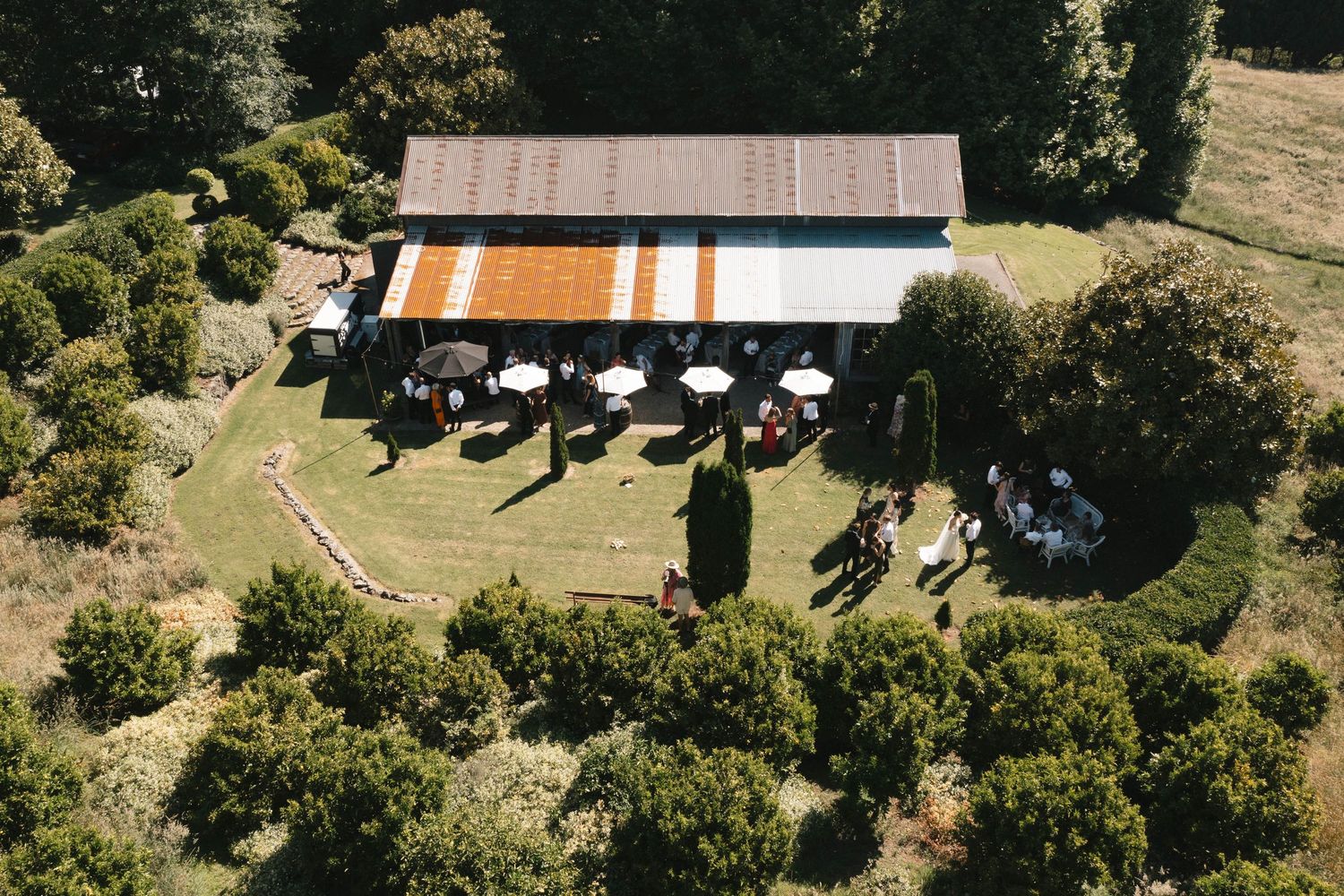 Aerial view of a rustic barn venue with outdoor seating area surrounded by lush green trees and landscaping.