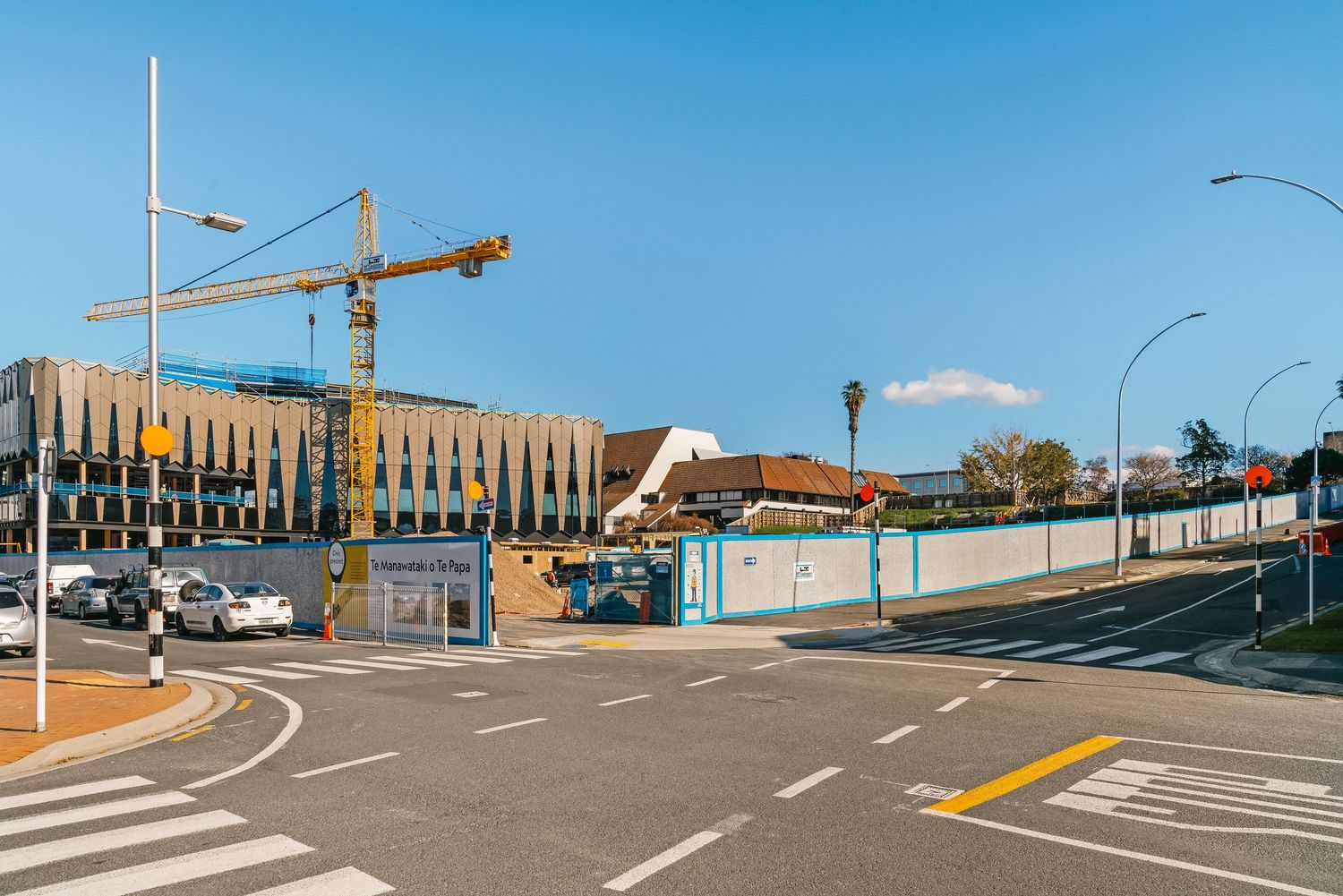 Construction cranes and walls line an empty curved road with yellow markings against a bright blue sky.