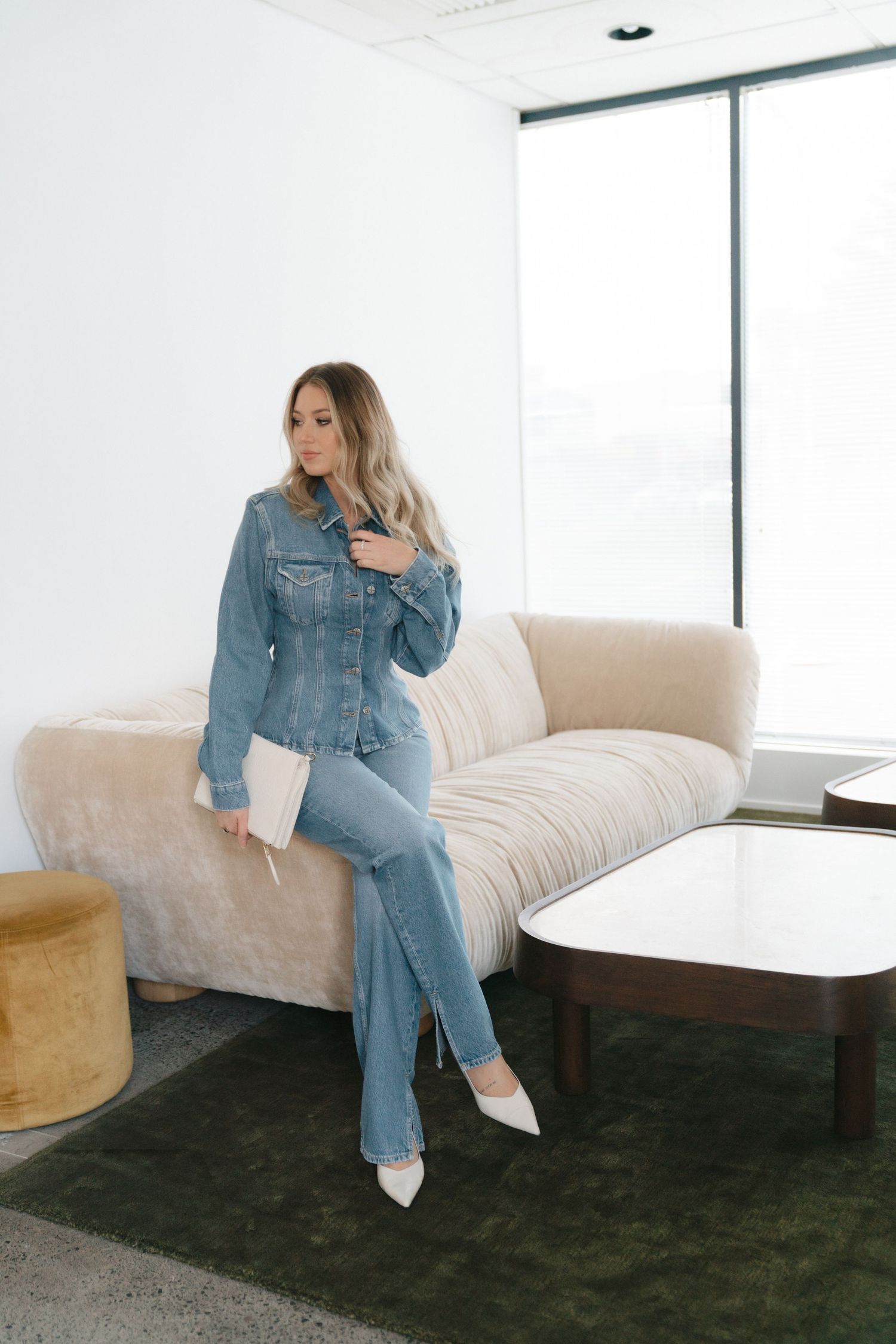 A denim-on-denim outfit paired with white heels while lounging on a cream couch in a minimalist living space.