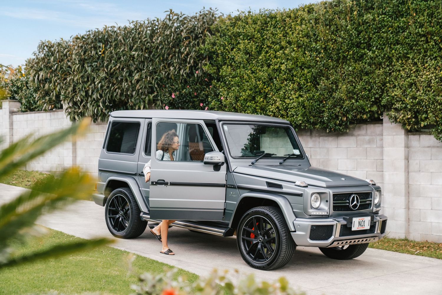 Silver Mercedes G-Wagon SUV parked in front of a lush green hedge on a sunny day.