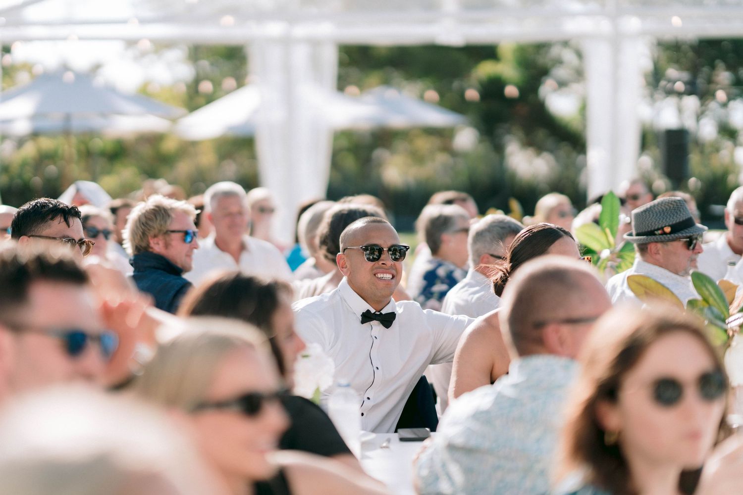 Wedding attendees seated in rows under bright sunlight wearing sunglasses and summer attire.