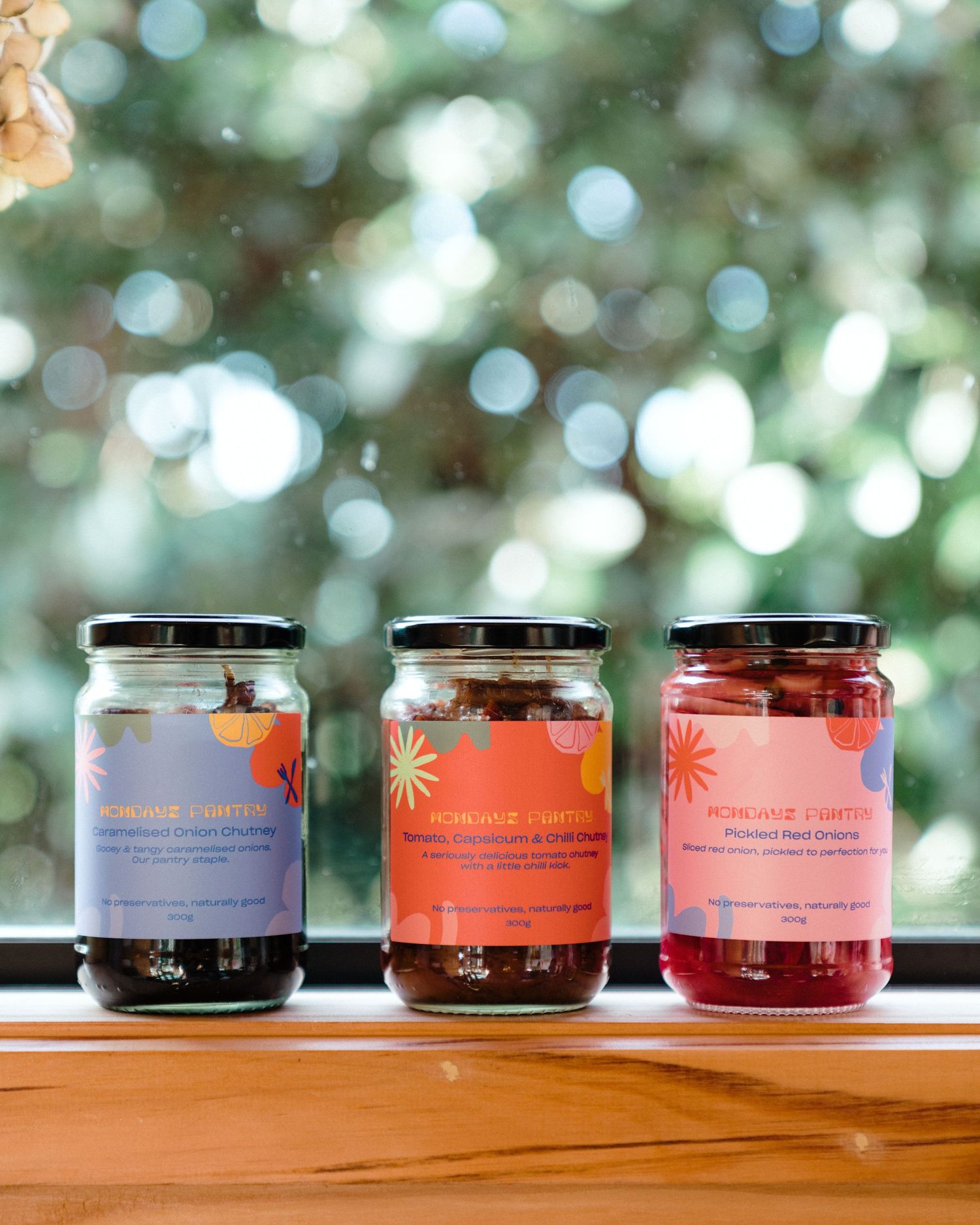 Three glass jars of colorful fruit preserves displayed on wooden ledge with bokeh background.