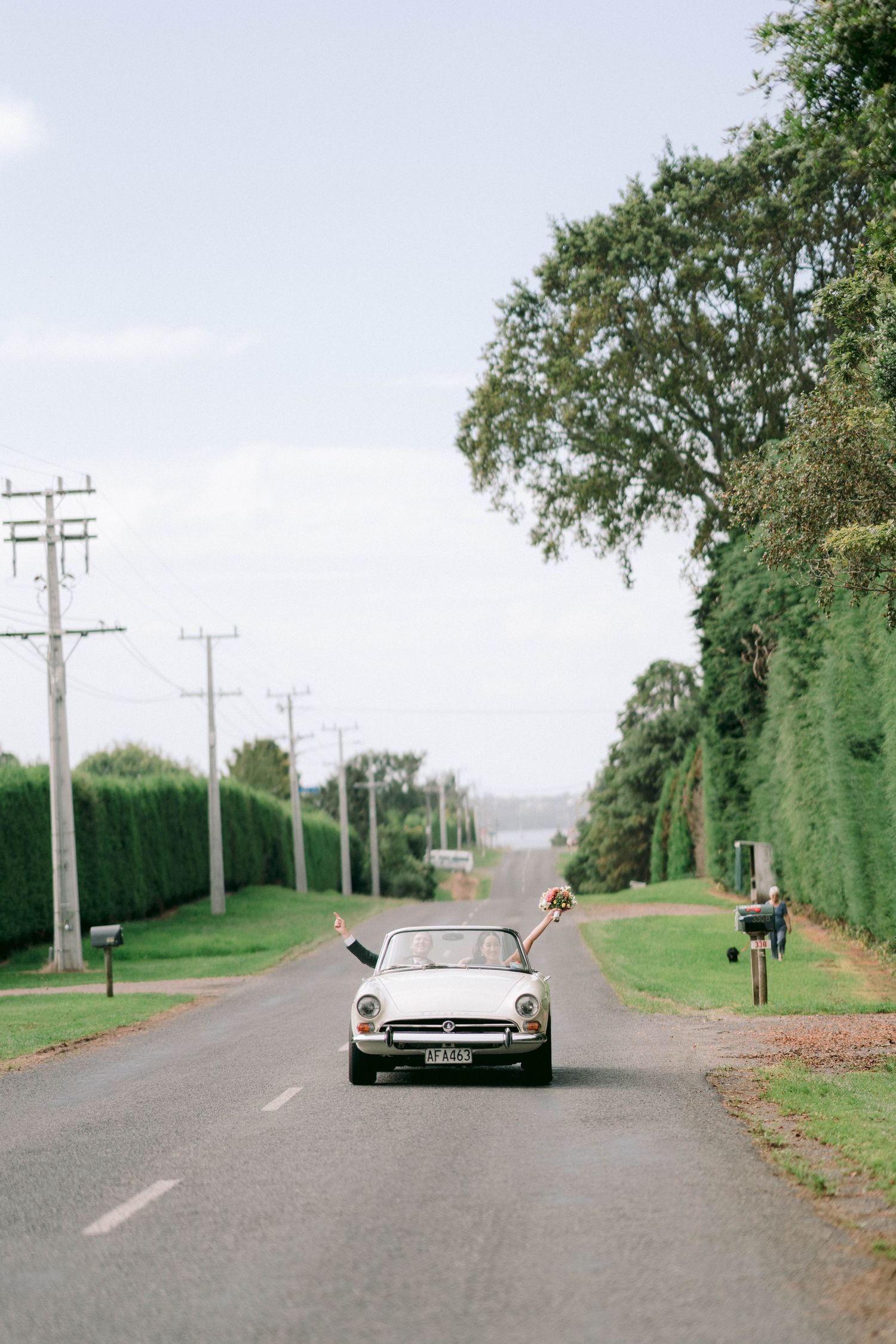 A classic convertible car drives down a tree-lined suburban road with neatly trimmed hedges on both sides.