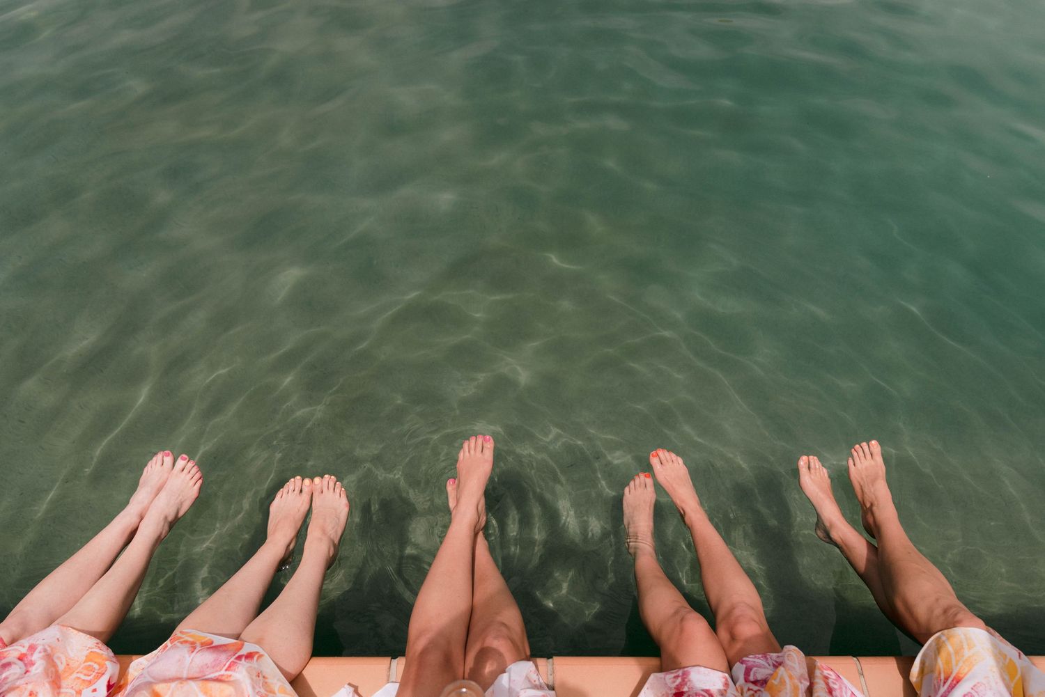 Feet and legs dangling over water's edge creating ripples in turquoise swimming pool.