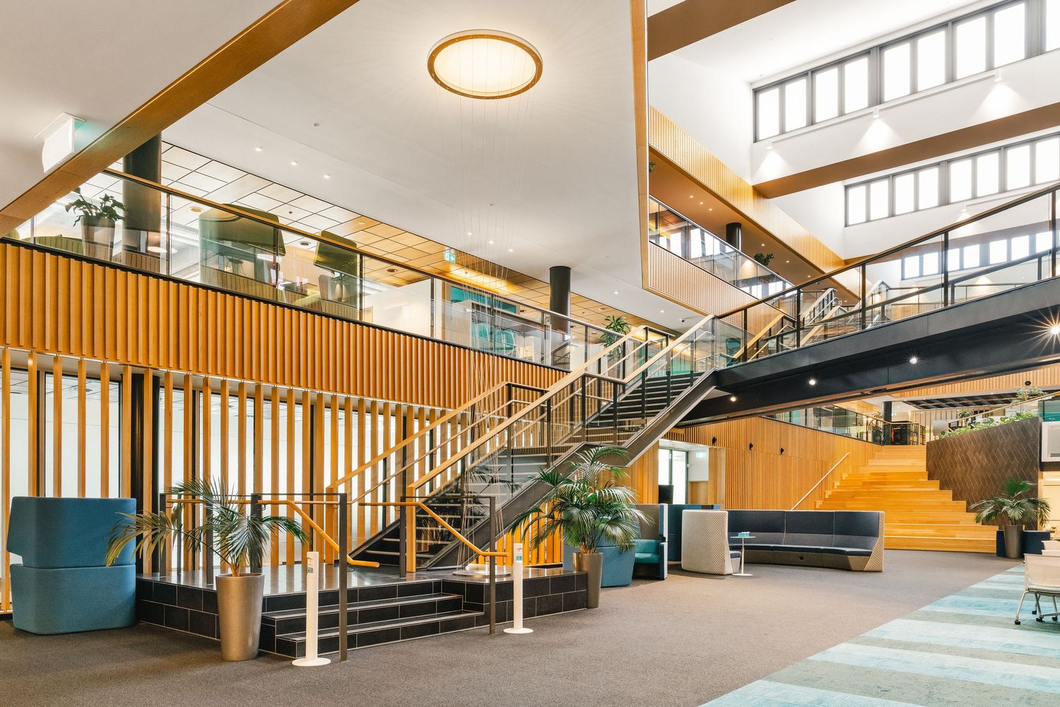 Modern office building interior featuring wood paneled walls, metal stairs, and yellow seating area with natural lighting.