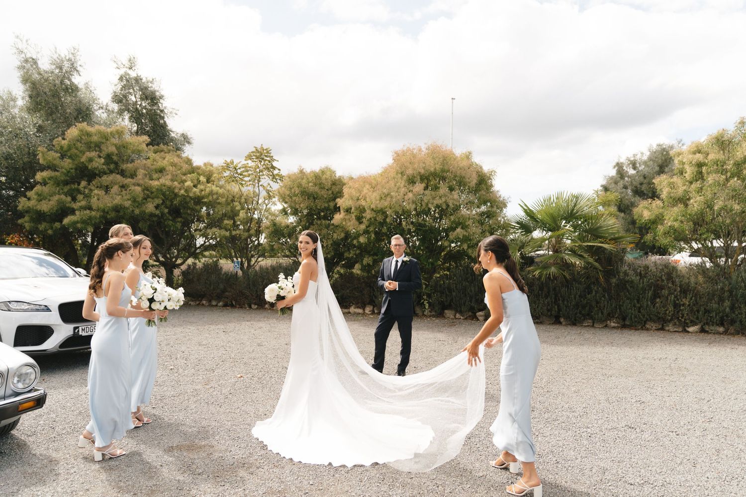 Bridesmaids help adjust a flowing white wedding gown train in an outdoor parking area on a cloudy day.