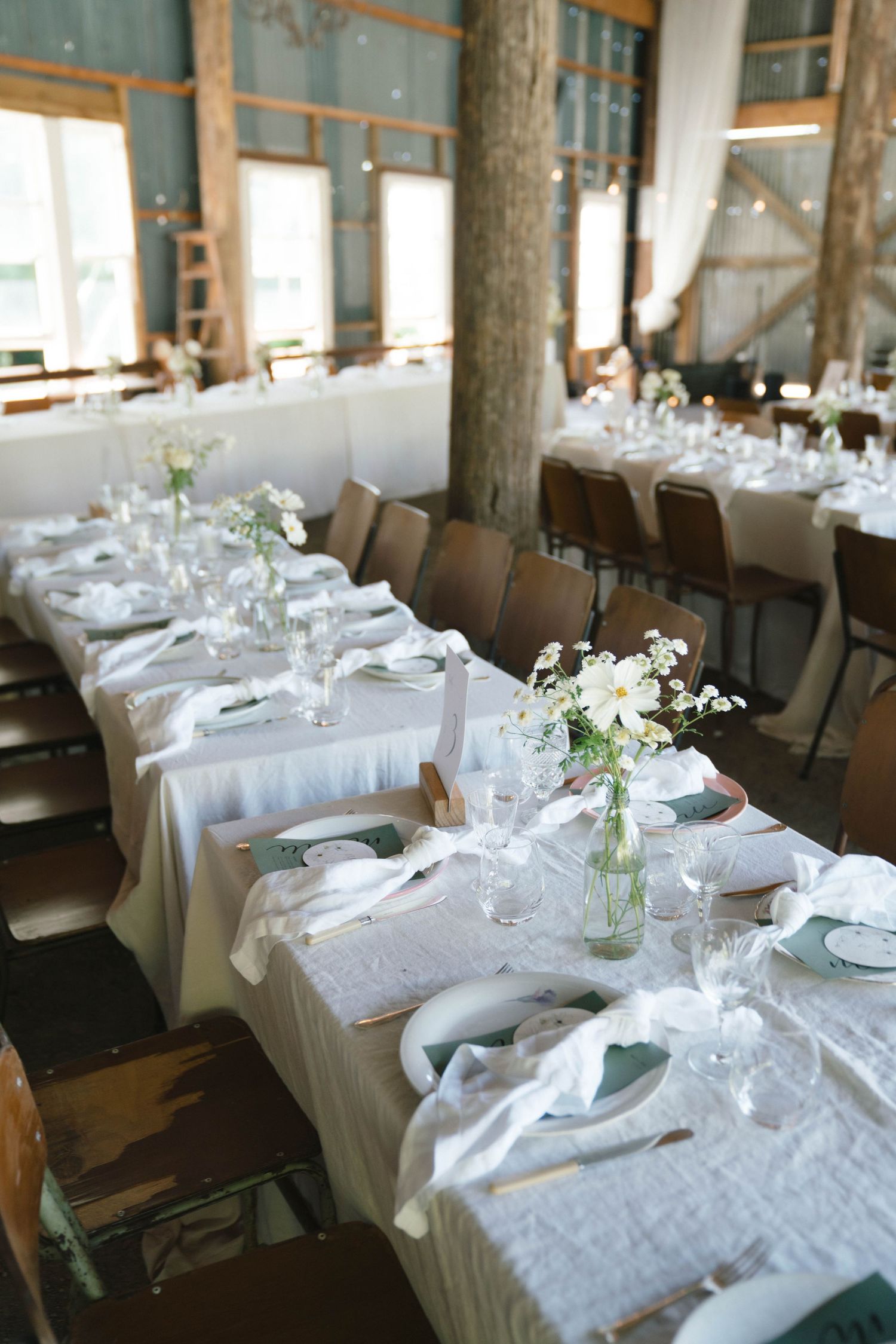 Long tables set with white linens and simple floral centerpieces in a rustic venue with exposed tree trunks.