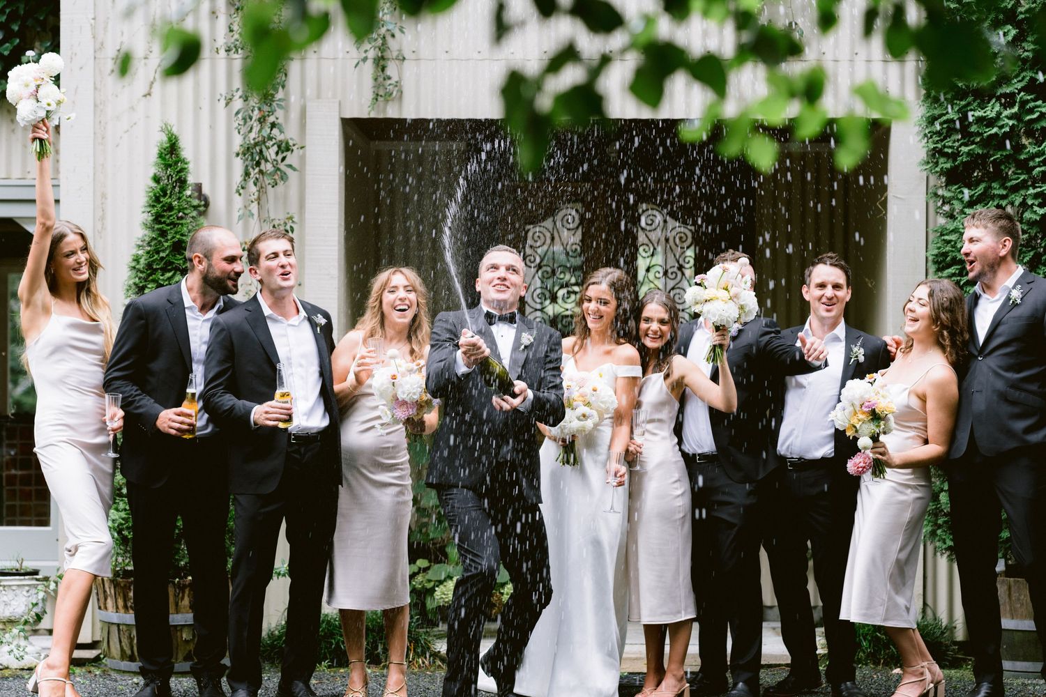 Wedding party pops champagne bottles outdoors against a white wall backdrop.