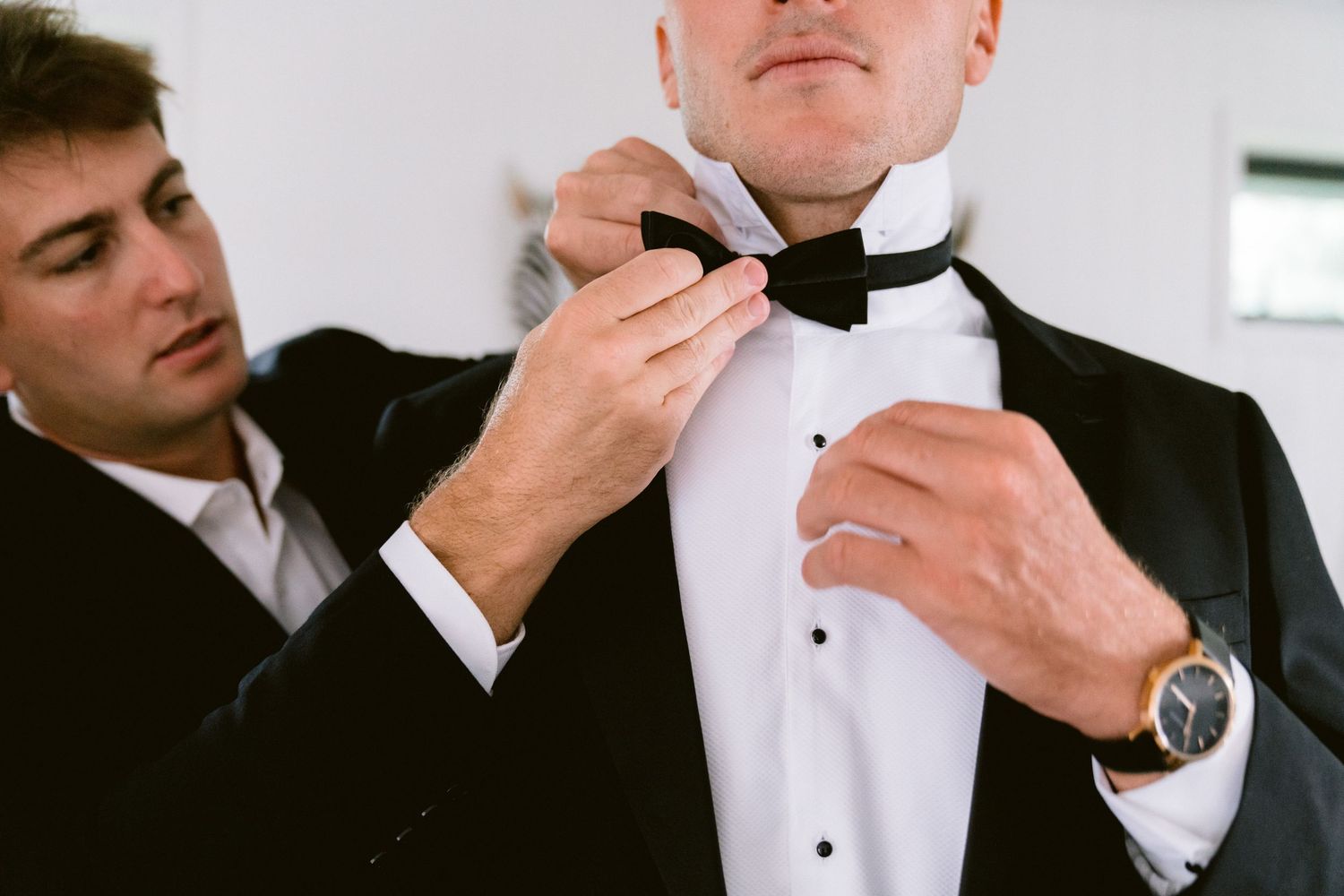 A man in a formal black tuxedo puts on a wristwatch while getting dressed.