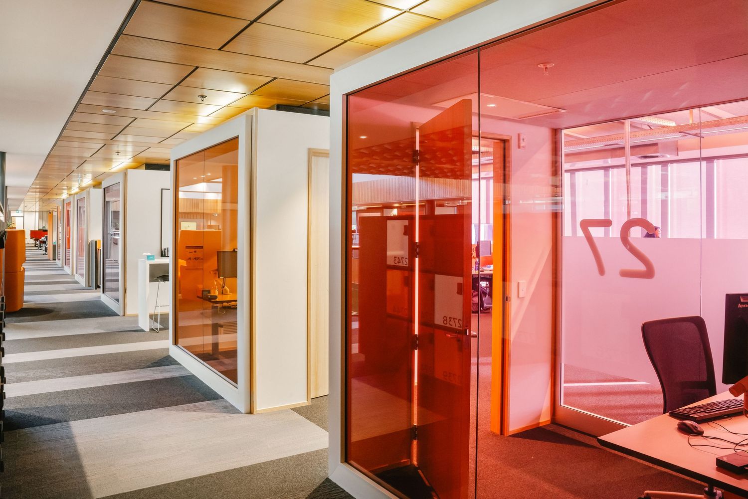 Modern office corridor with glass meeting rooms featuring vibrant red-tinted partitions and warm overhead lighting.