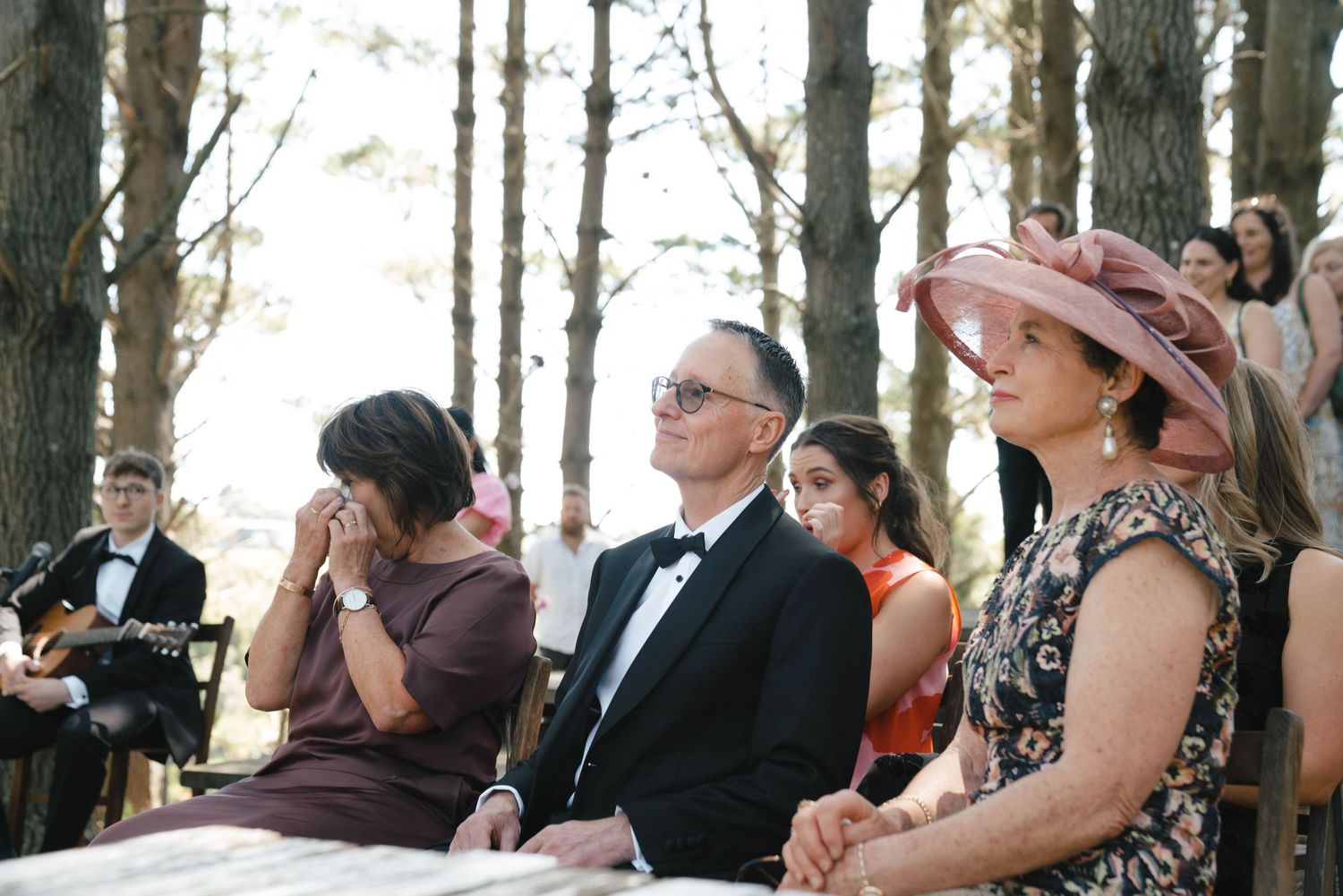 Guests in formal wear seated on wooden benches during an outdoor ceremony.