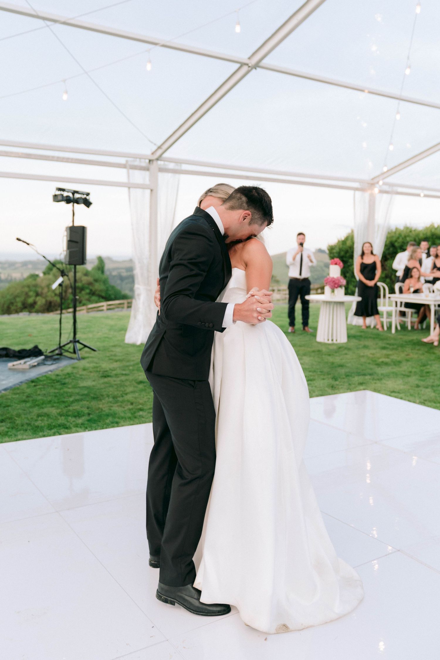Newlyweds share a romantic moment on white dance floor under elegant tent with scenic views.