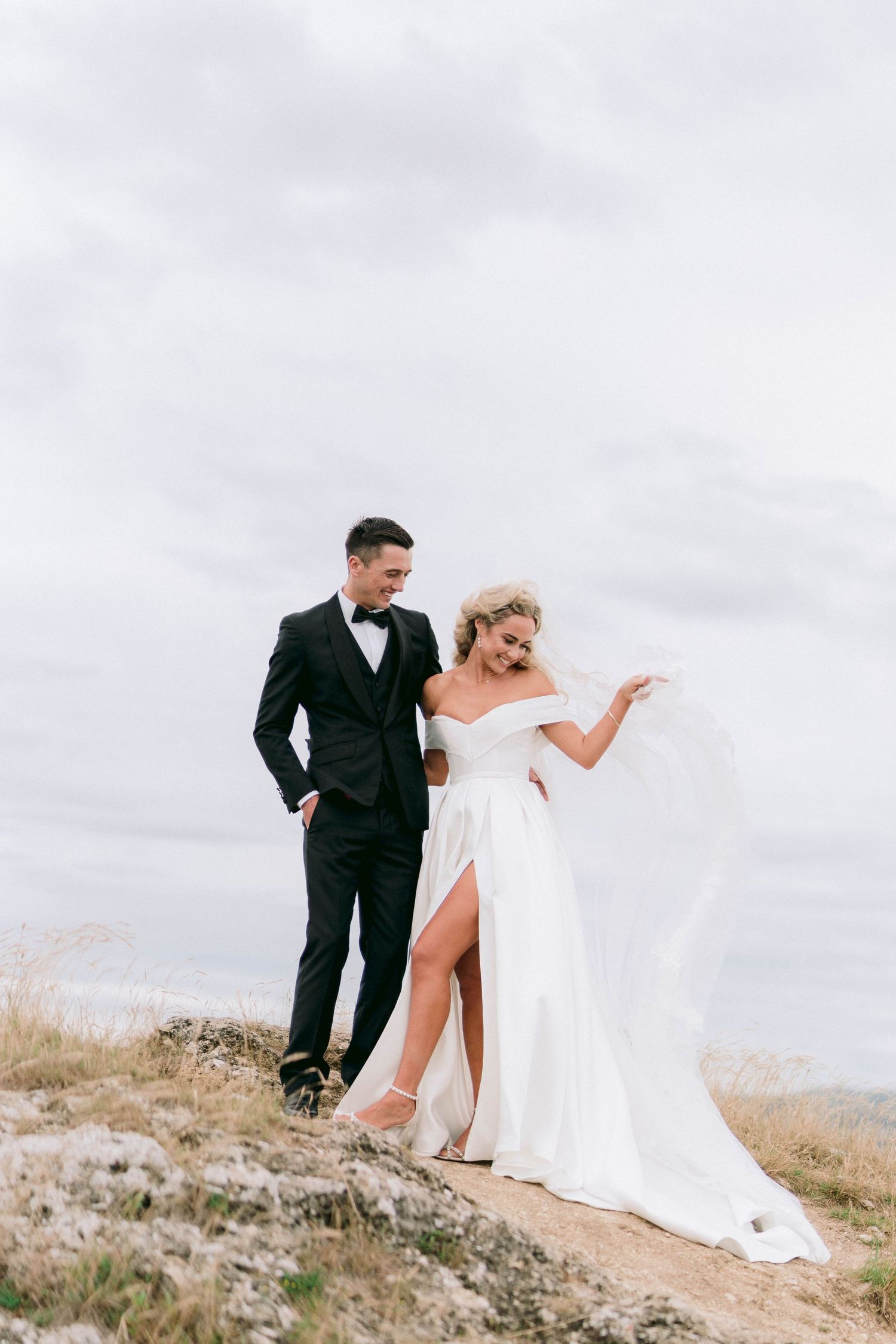 A newlywed couple poses dramatically on a hilltop, with the bride wearing a flowing white gown with a high slit.