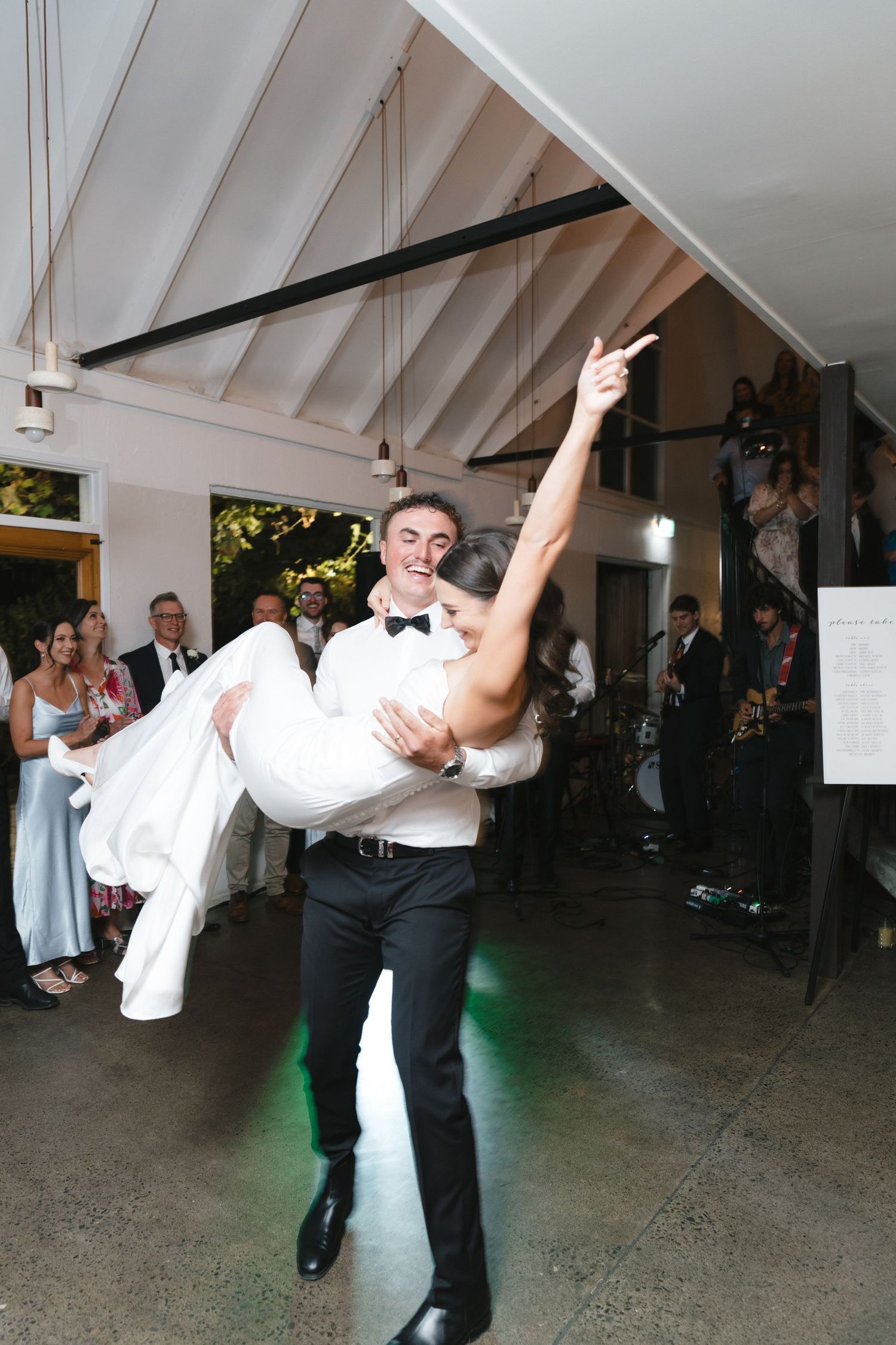 Groom carrying bride during their first dance in a rustic venue with exposed beam ceiling.