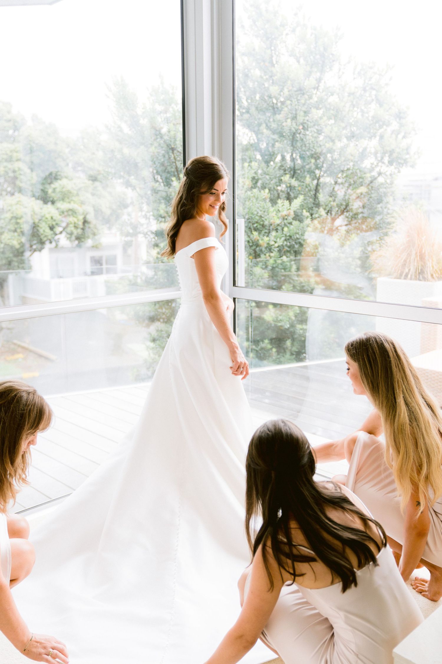 Bridesmaids in white dresses gather by large windows during wedding preparations in a bright room.