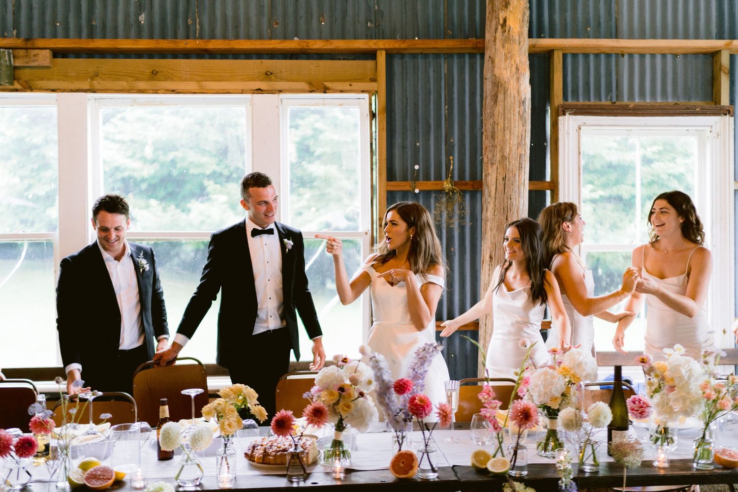 Wedding party gathered at a rustic reception table decorated with colorful floral centerpieces.