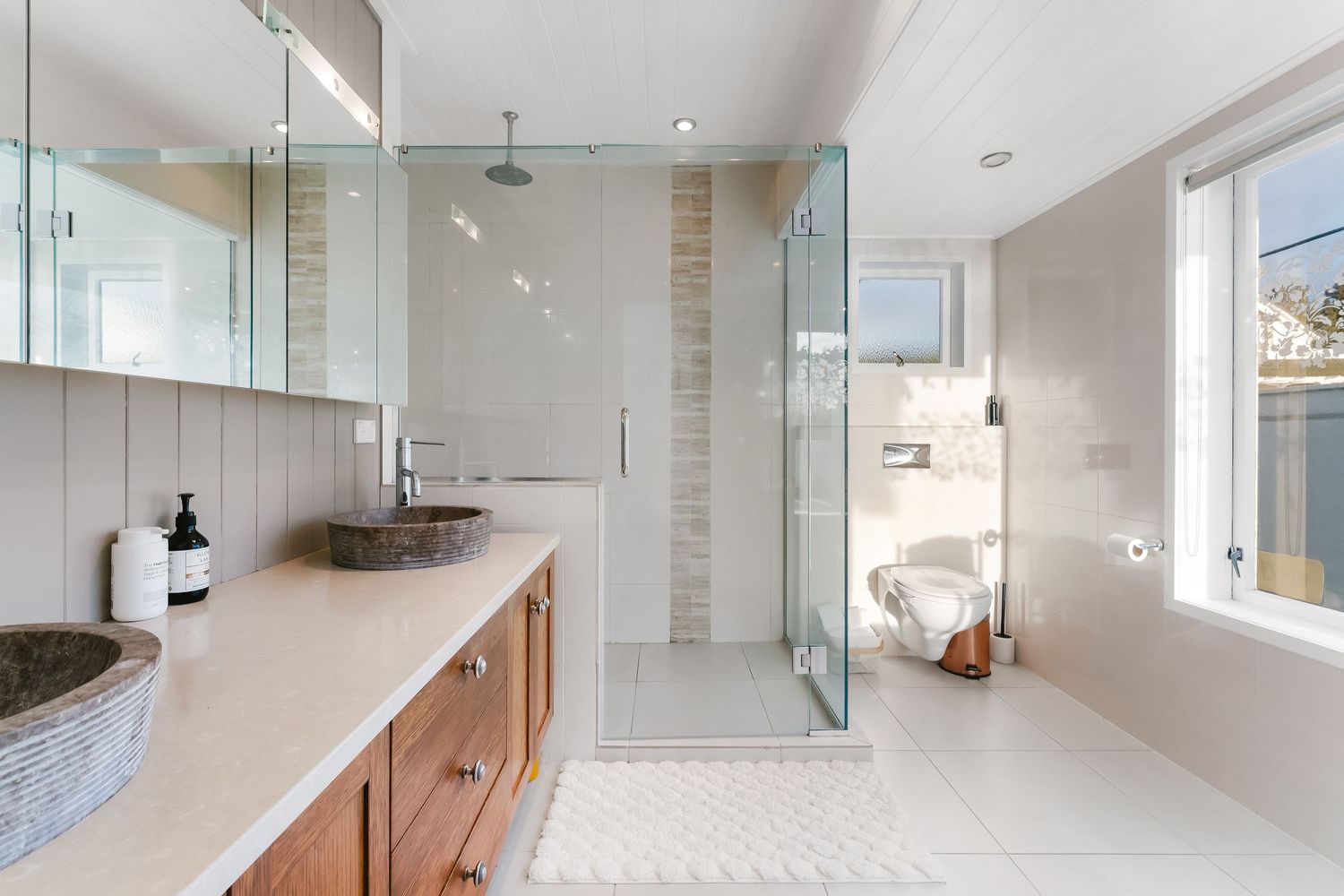 Modern bathroom with stone vessel sinks, wooden vanity, glass shower enclosure and natural light from window.