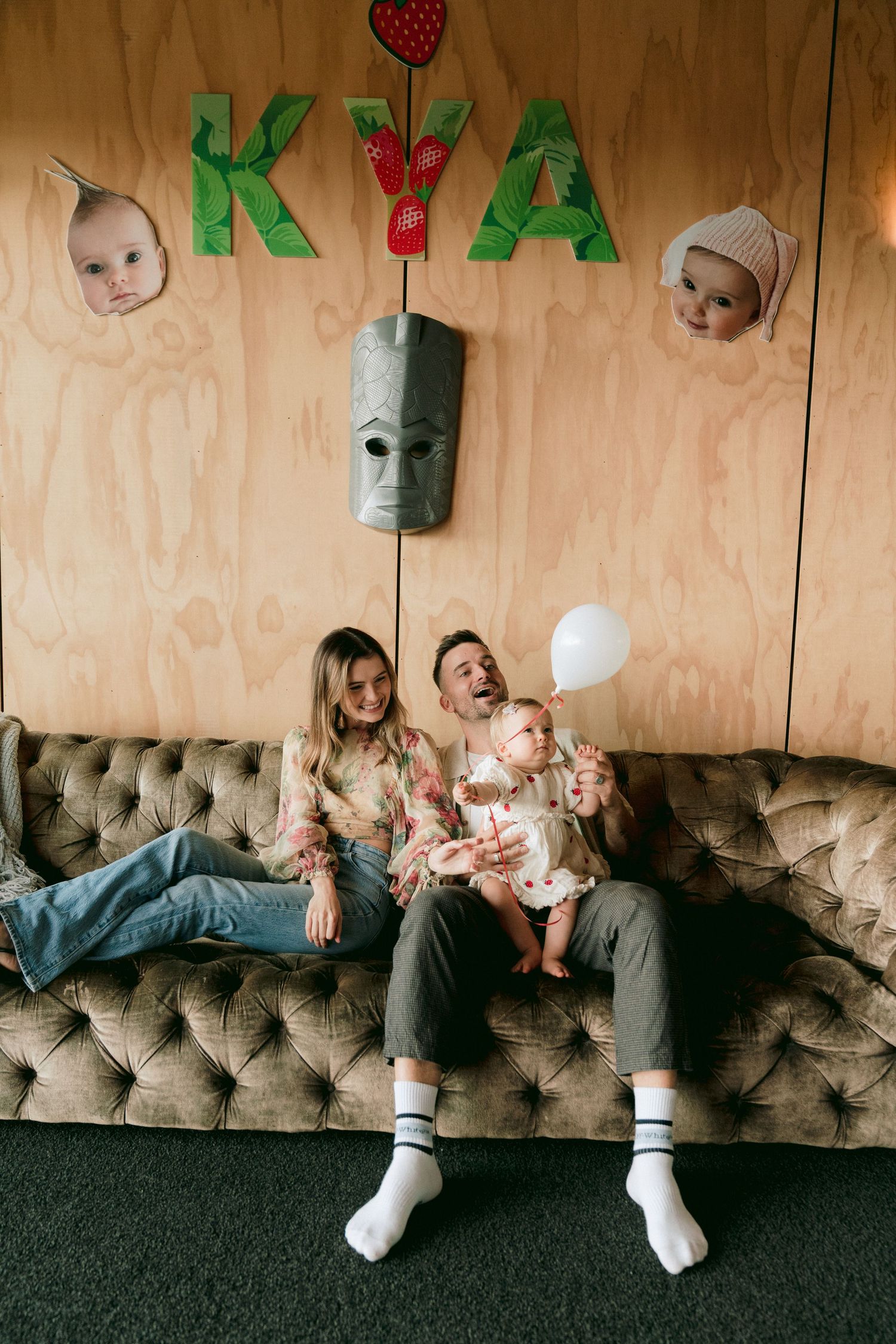 Family sitting on a vintage velvet couch under decorative letters and balloon in a casual indoor setting.