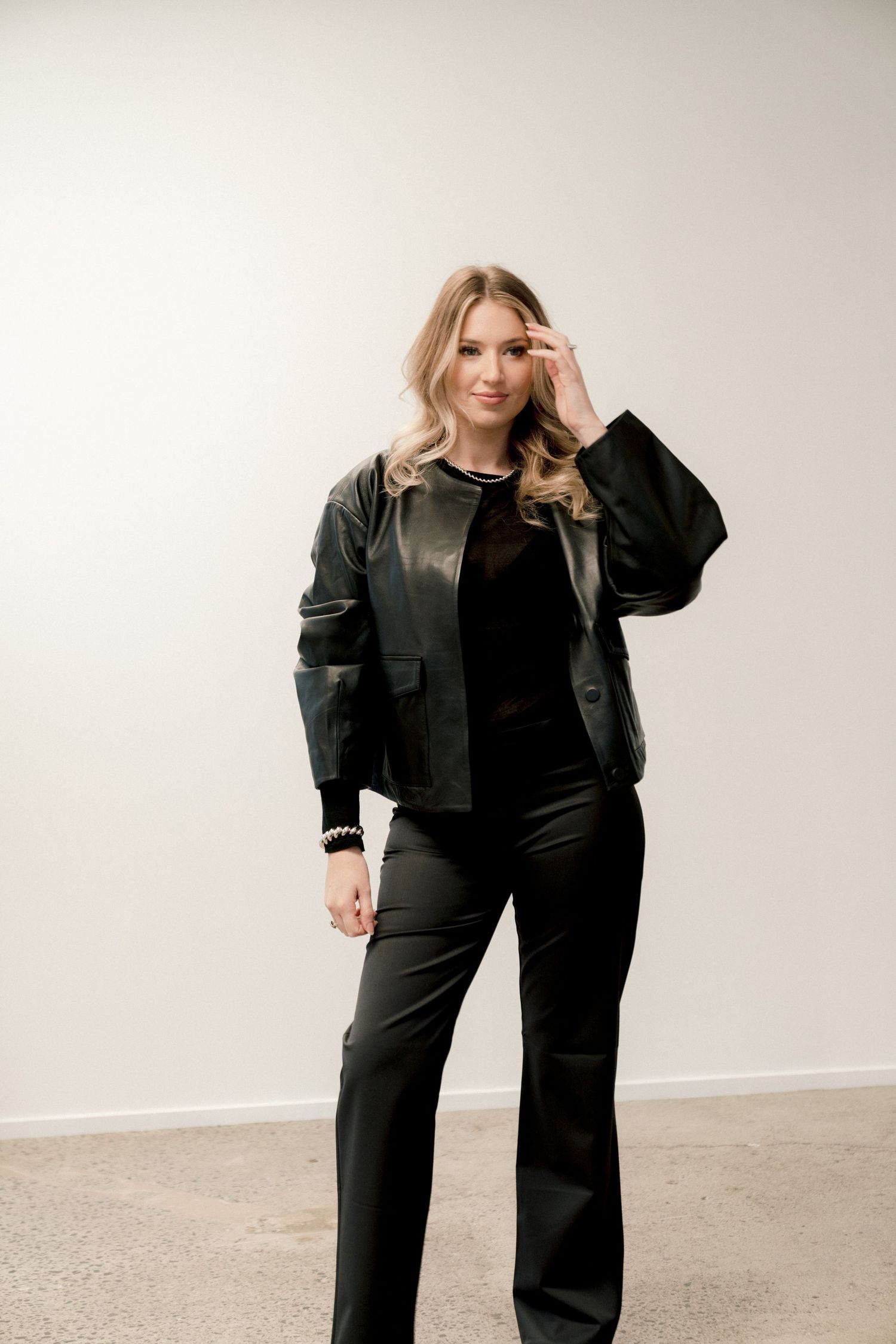 A person wearing a black leather jacket and wide-leg pants poses casually against a white studio backdrop.