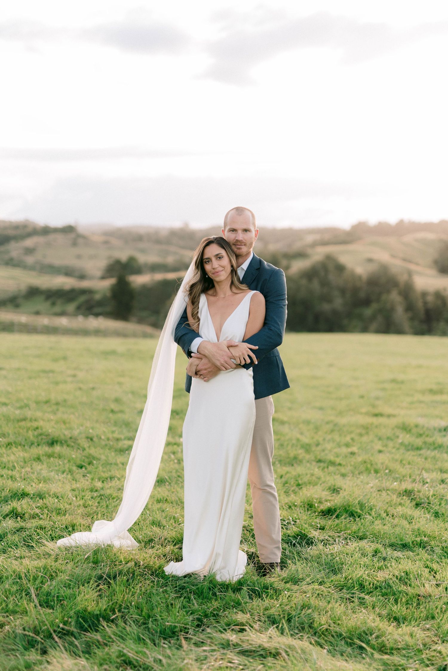 A bride in white dress and groom in blue jacket pose playfully in a grassy field.