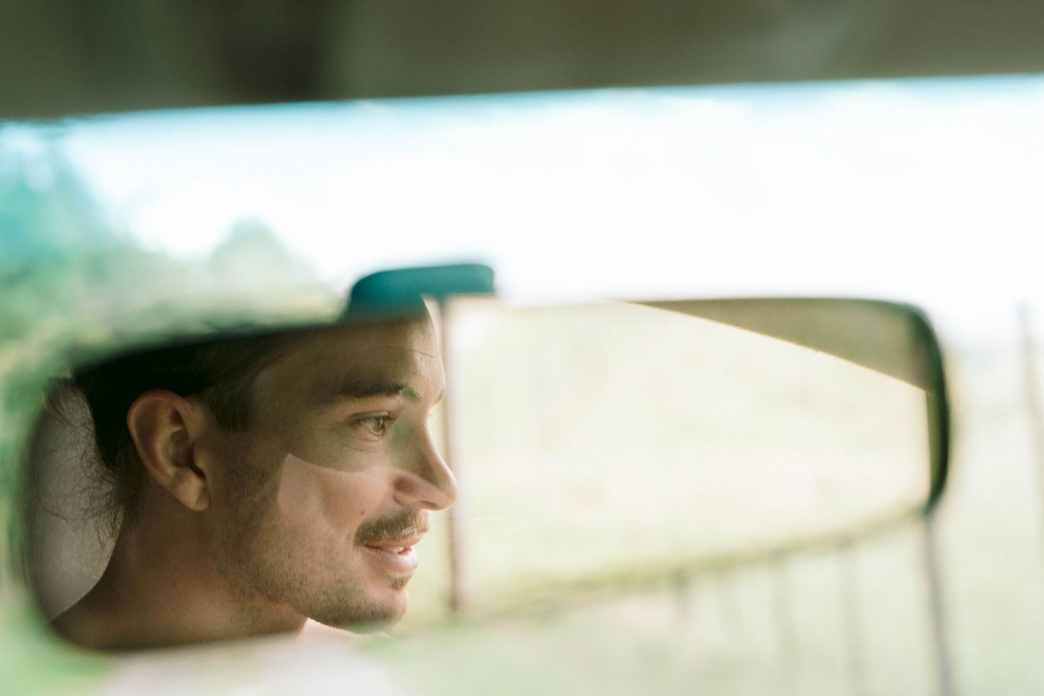 A rearview mirror reflection shows a profile view of a person behind the wheel of a car on a bright sunny day.