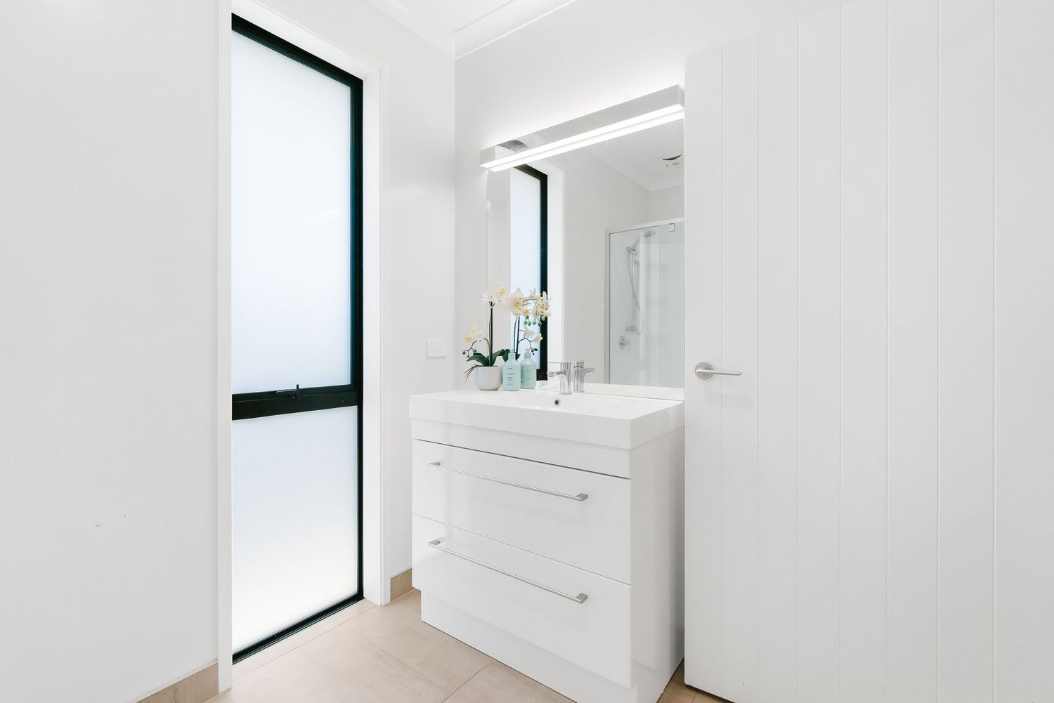 Modern white bathroom vanity with large mirror, sleek drawers and tall frosted window in minimalist style.