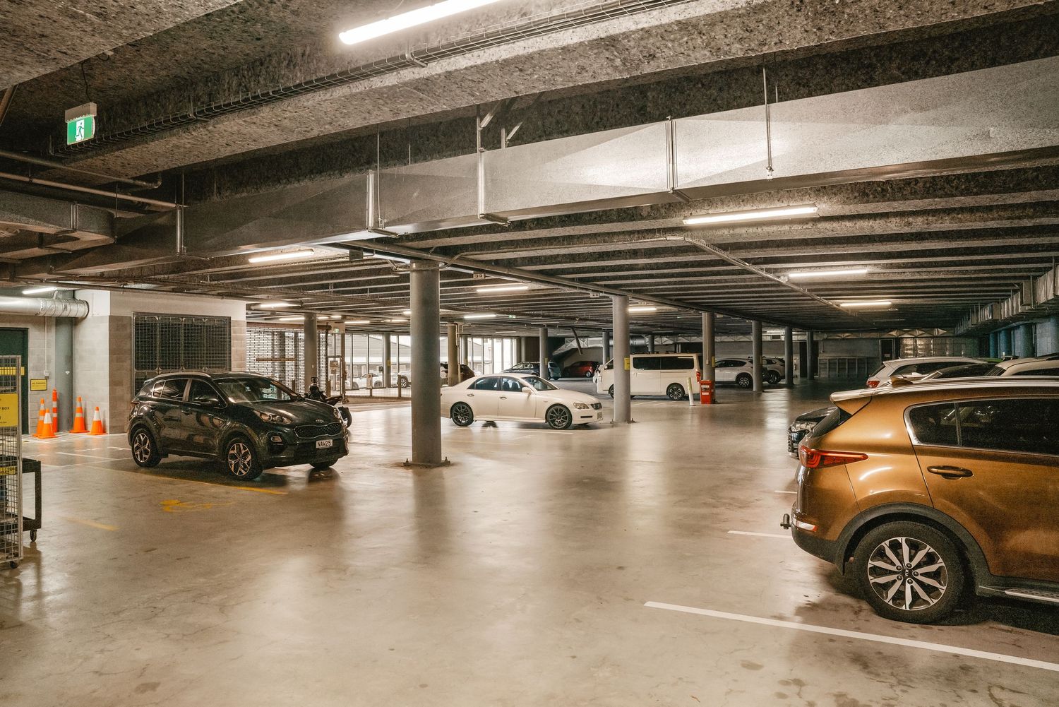 Modern indoor parking garage with concrete columns and various parked vehicles under fluorescent lighting.