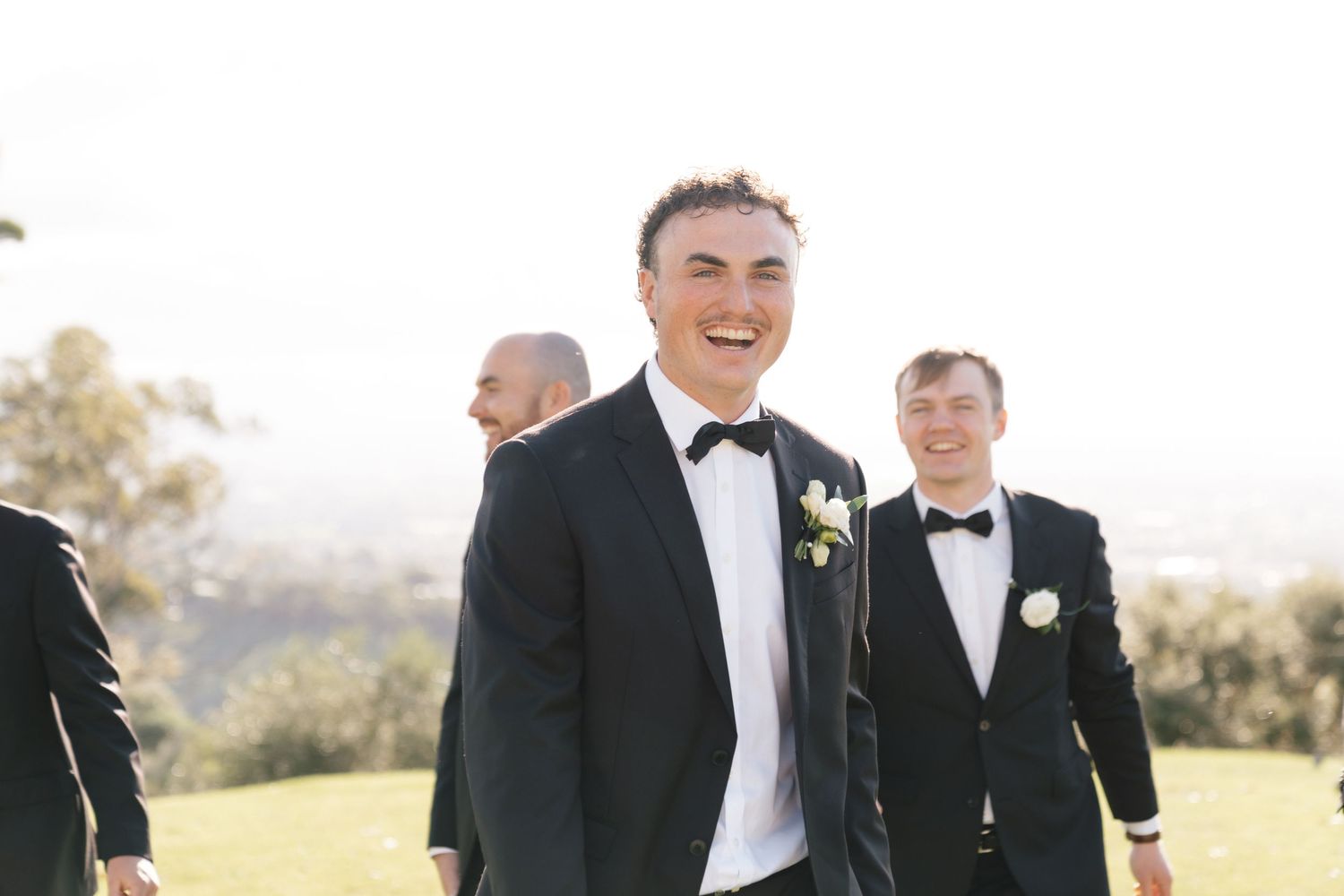 Wedding party in black formal attire poses outdoors during golden hour at scenic overlook.