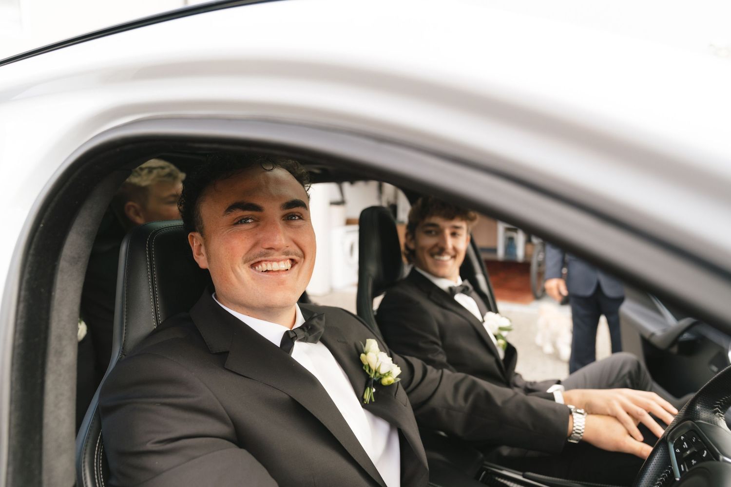 A person wearing a tuxedo with boutonniere sits in car smiling while driving during a formal event.