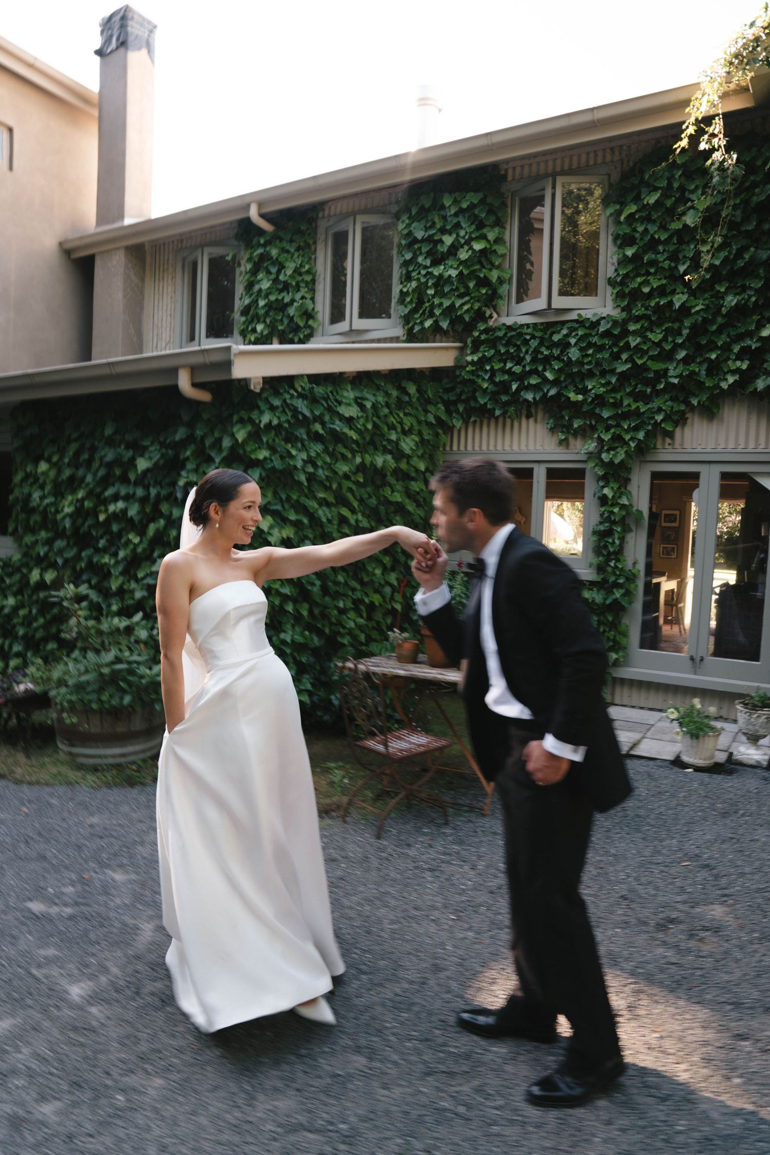 A couple in formal wedding attire playfully interacting in front of an ivy-covered building during their special day.