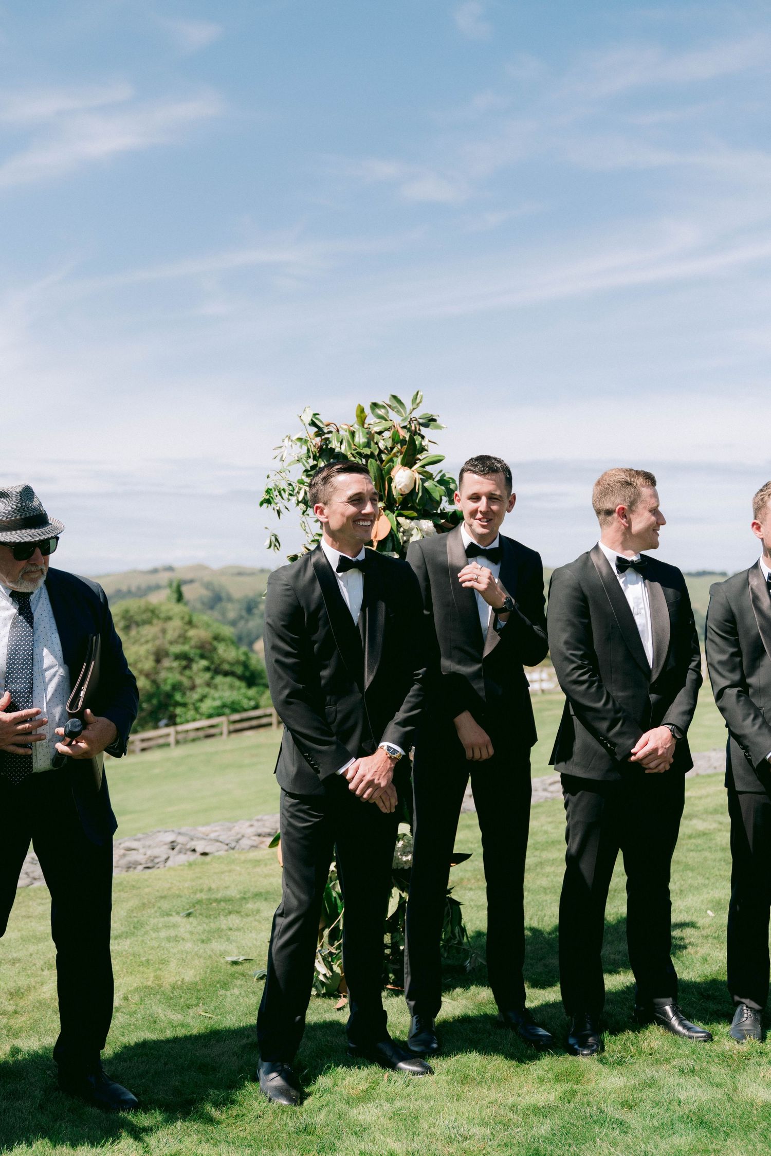 A line of groomsmen dressed in black suits enjoying a playful moment on a green hillside under blue skies.