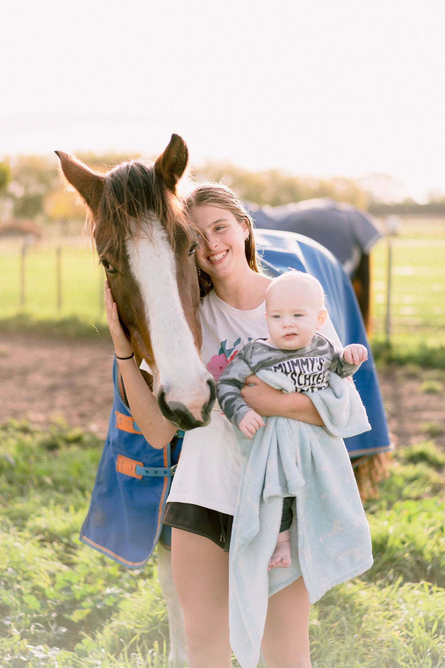 A brown and white horse nuzzles close to a young baby on a sunny day in a grassy field.