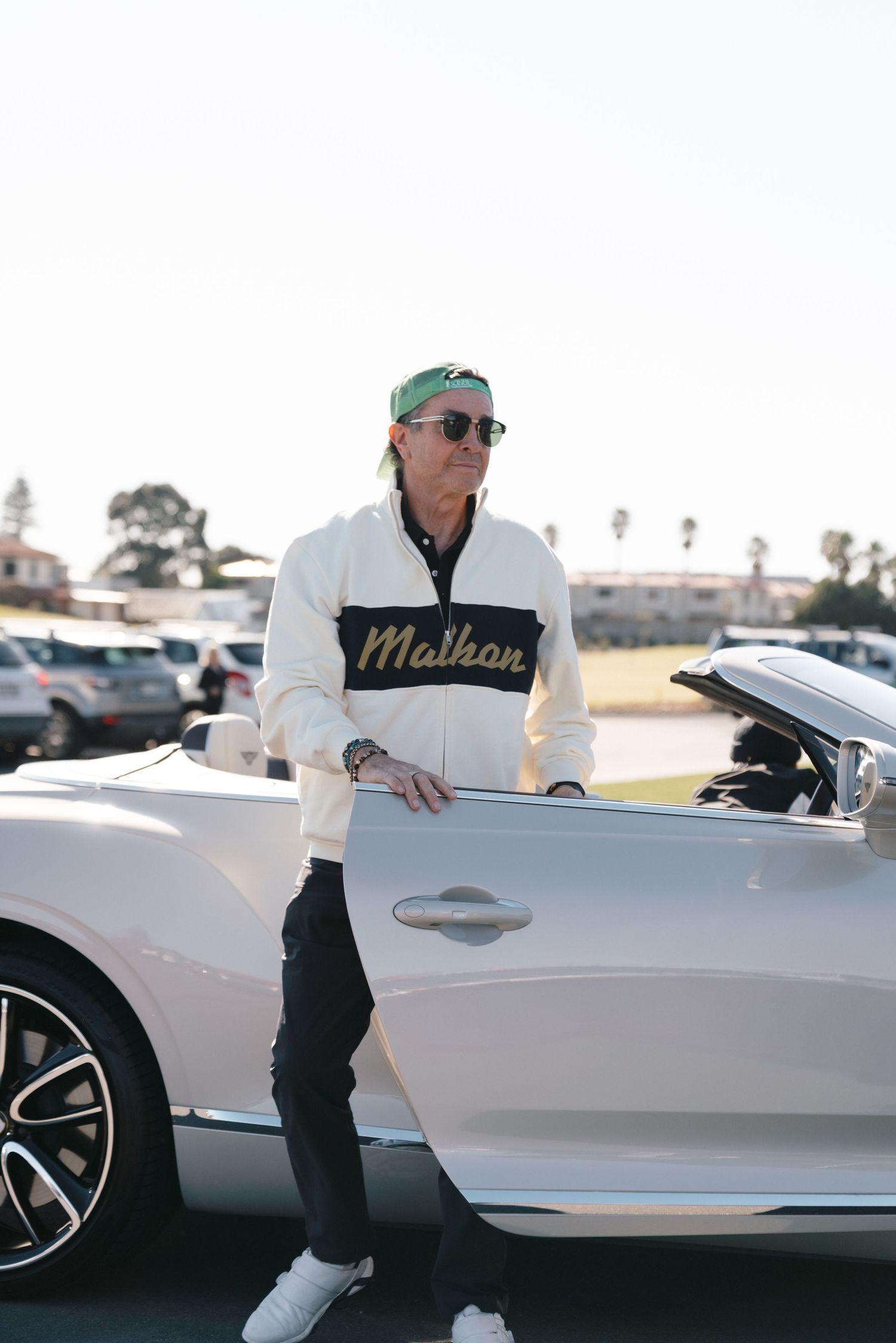 Person in white sweater and sunglasses stands beside white luxury convertible in parking lot.