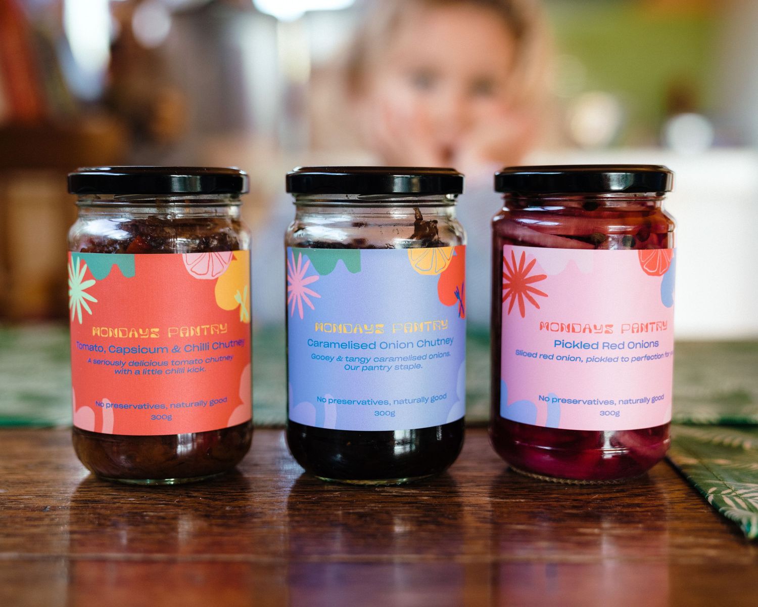 Three glass jars of colorful jam with pink labels sit on a wooden table.