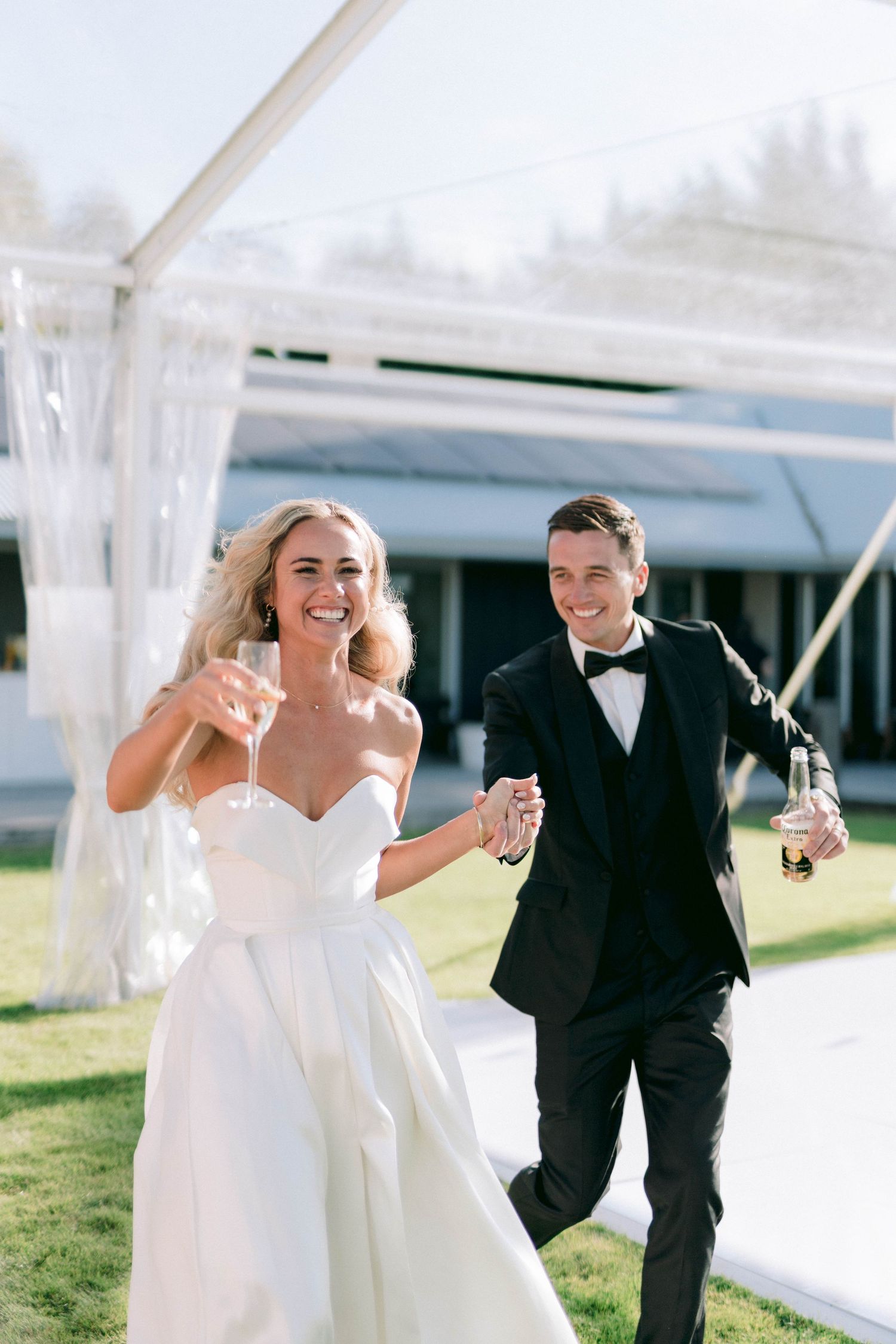 A joyful wedding couple toasting with champagne glasses while walking on grass under a tent.