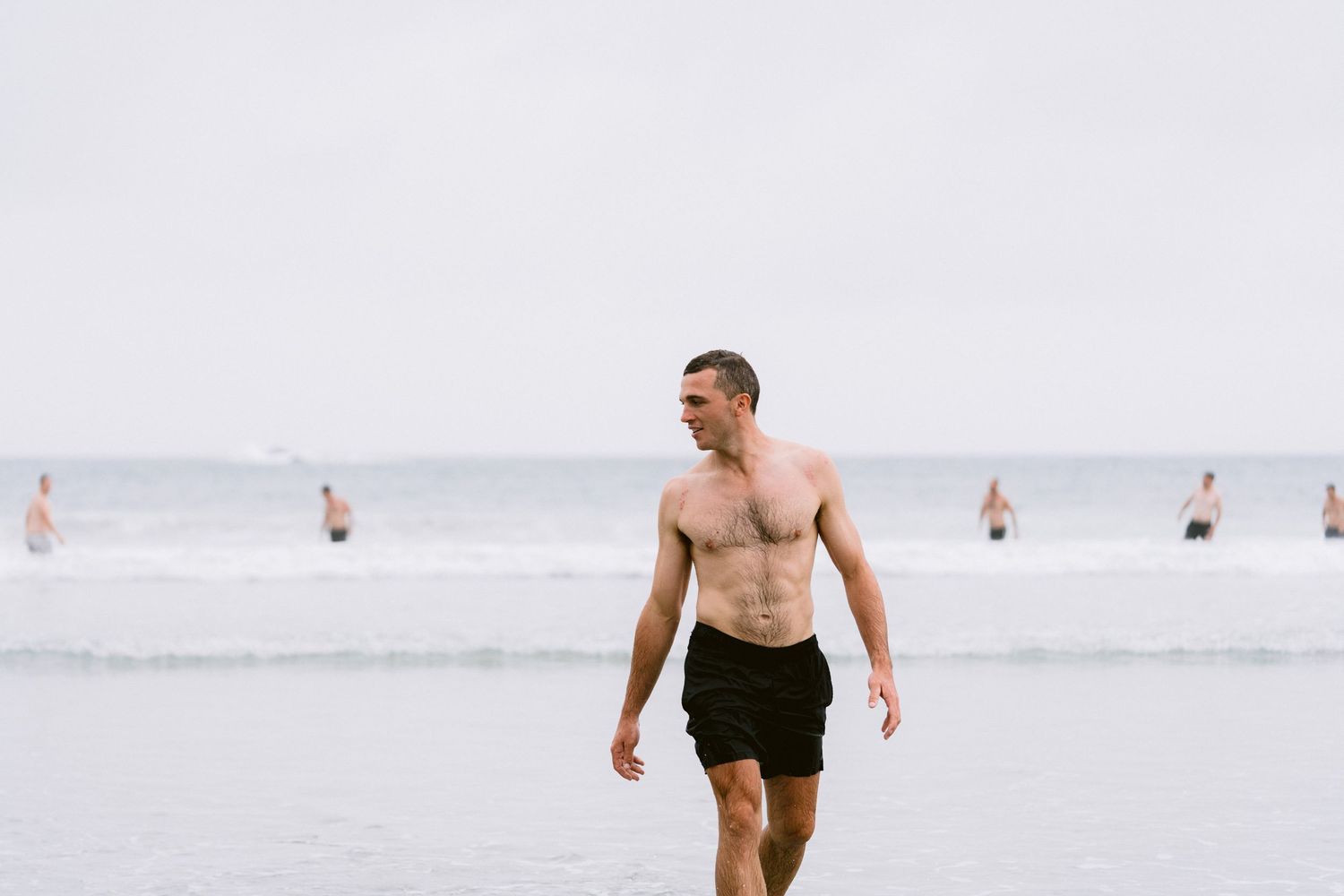A shirtless person in black shorts walks along a misty beach with people visible in the background.