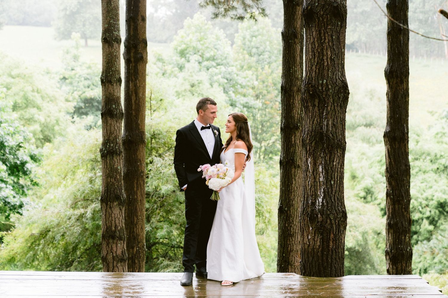 A wedding couple poses for romantic portraits under tall trees in a wooded outdoor setting.