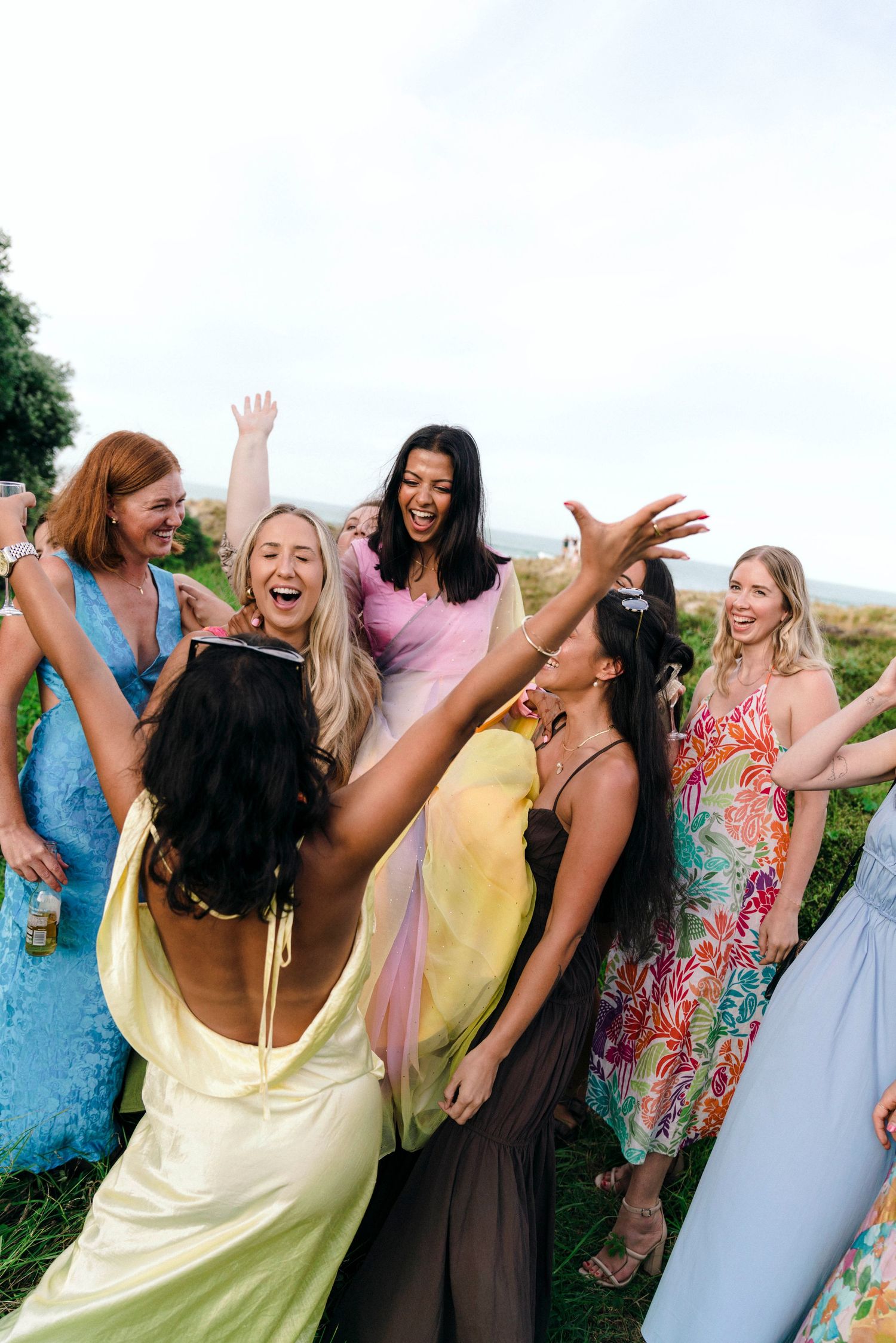 A group of friends in colorful summer dresses laughing and celebrating together outdoors on a sunny day.