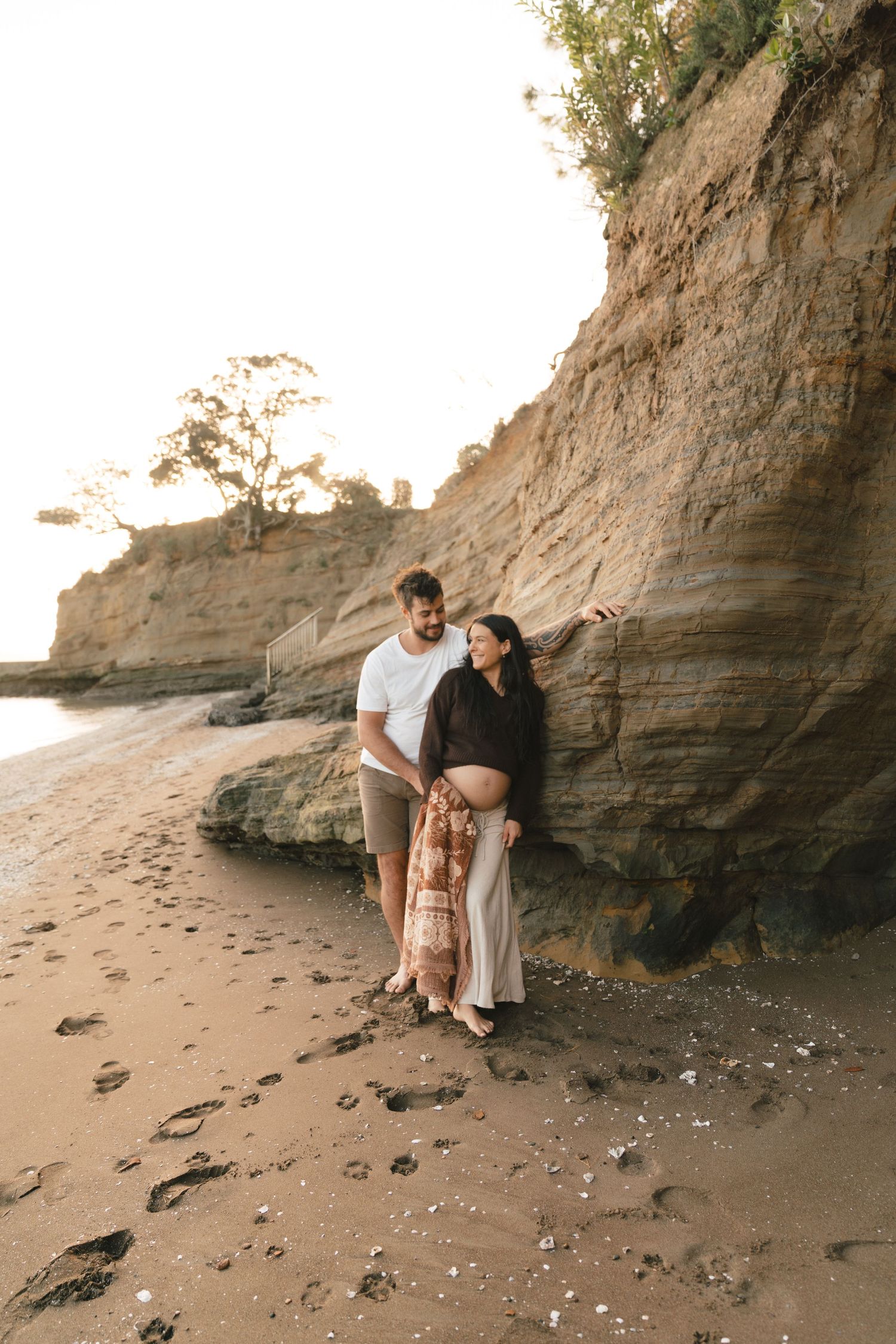 A couple shares an intimate moment against a rocky beach cliff during sunset, creating silhouettes in the warm light.