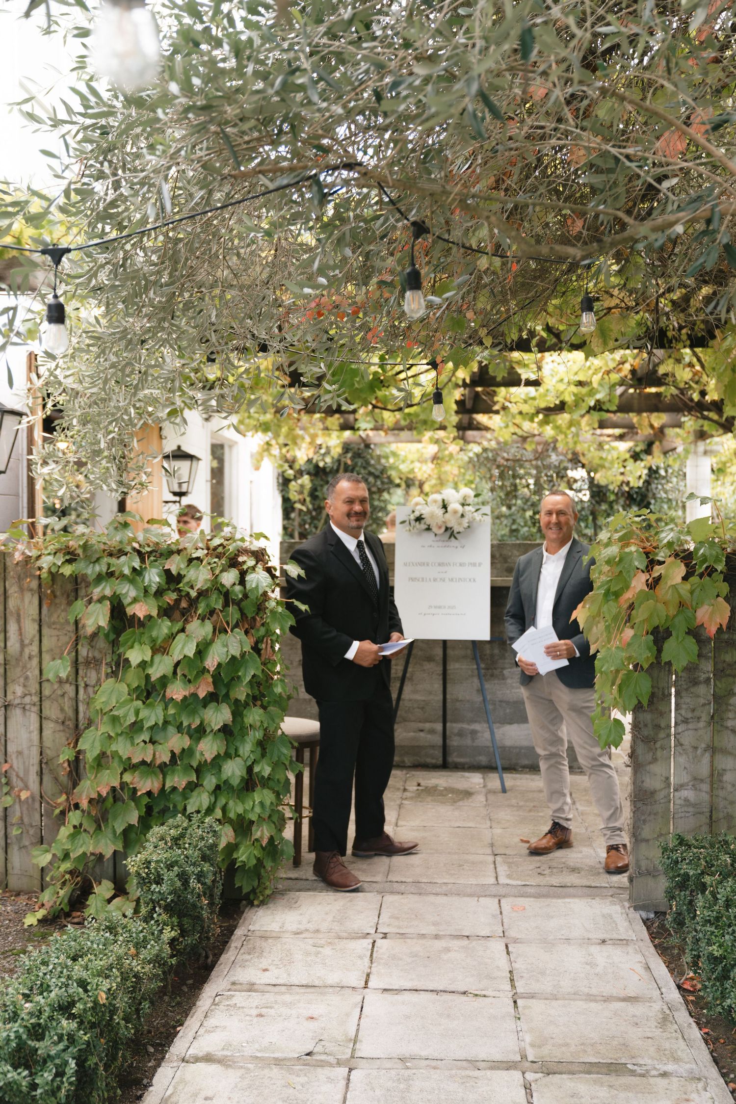 Two presenters stand beneath a vine-covered archway displaying information on an easel during an outdoor event.