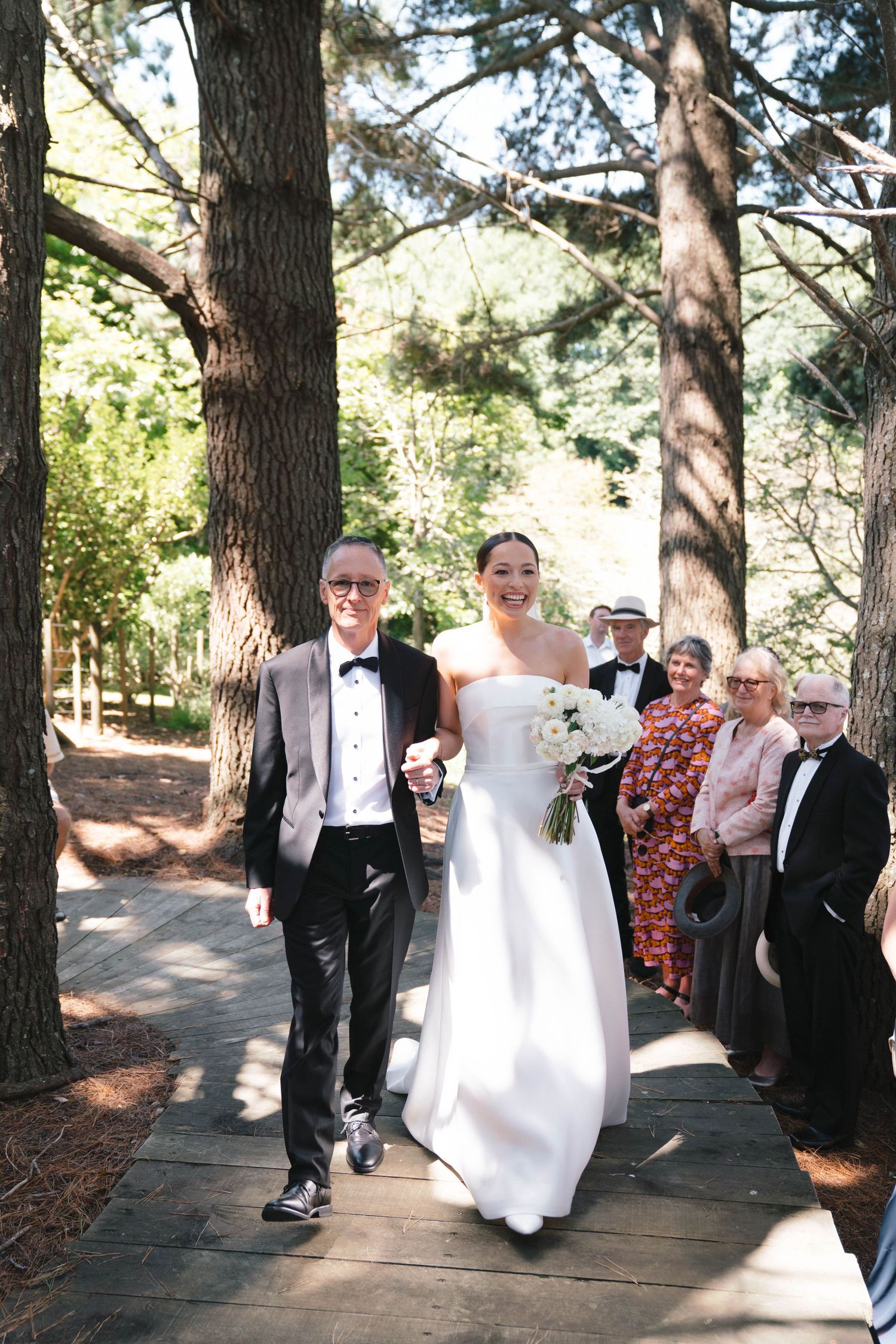 A wedding procession makes its way down a wooden walkway surrounded by pine trees.