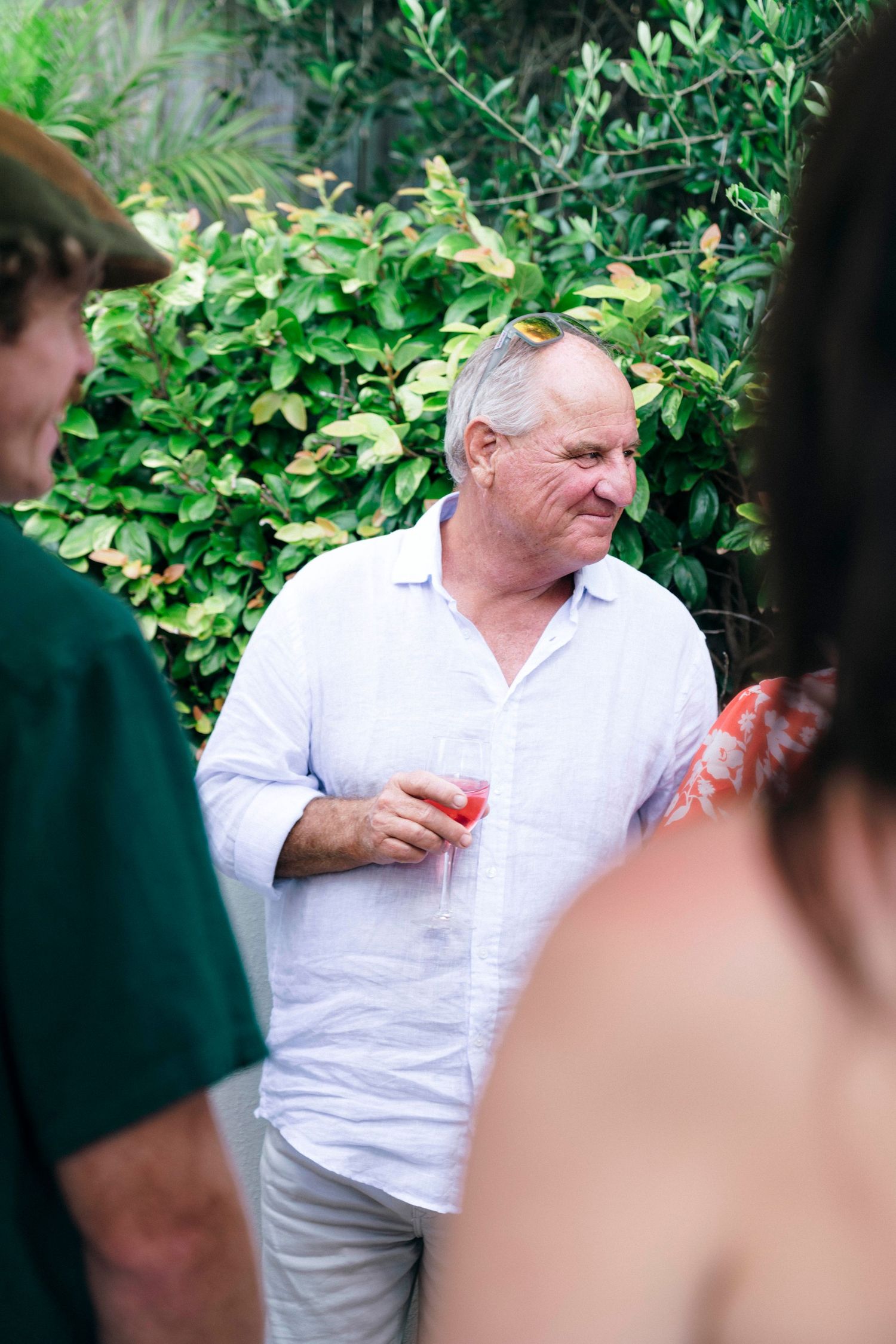 Man in white linen shirt and pants standing outdoors against green foliage backdrop during a social gathering.