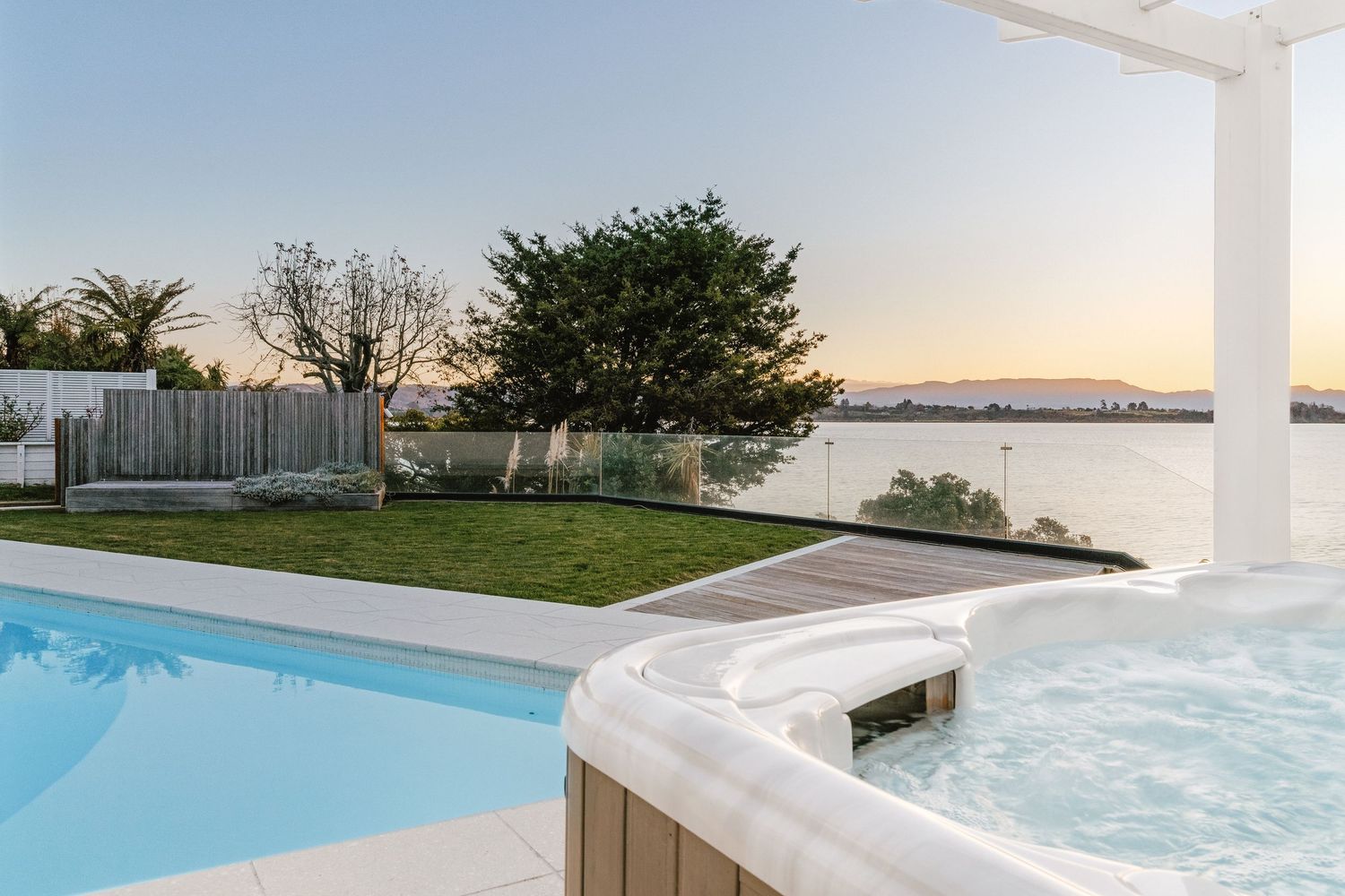 Luxurious backyard with infinity pool and hot tub overlooking waterfront at dusk.
