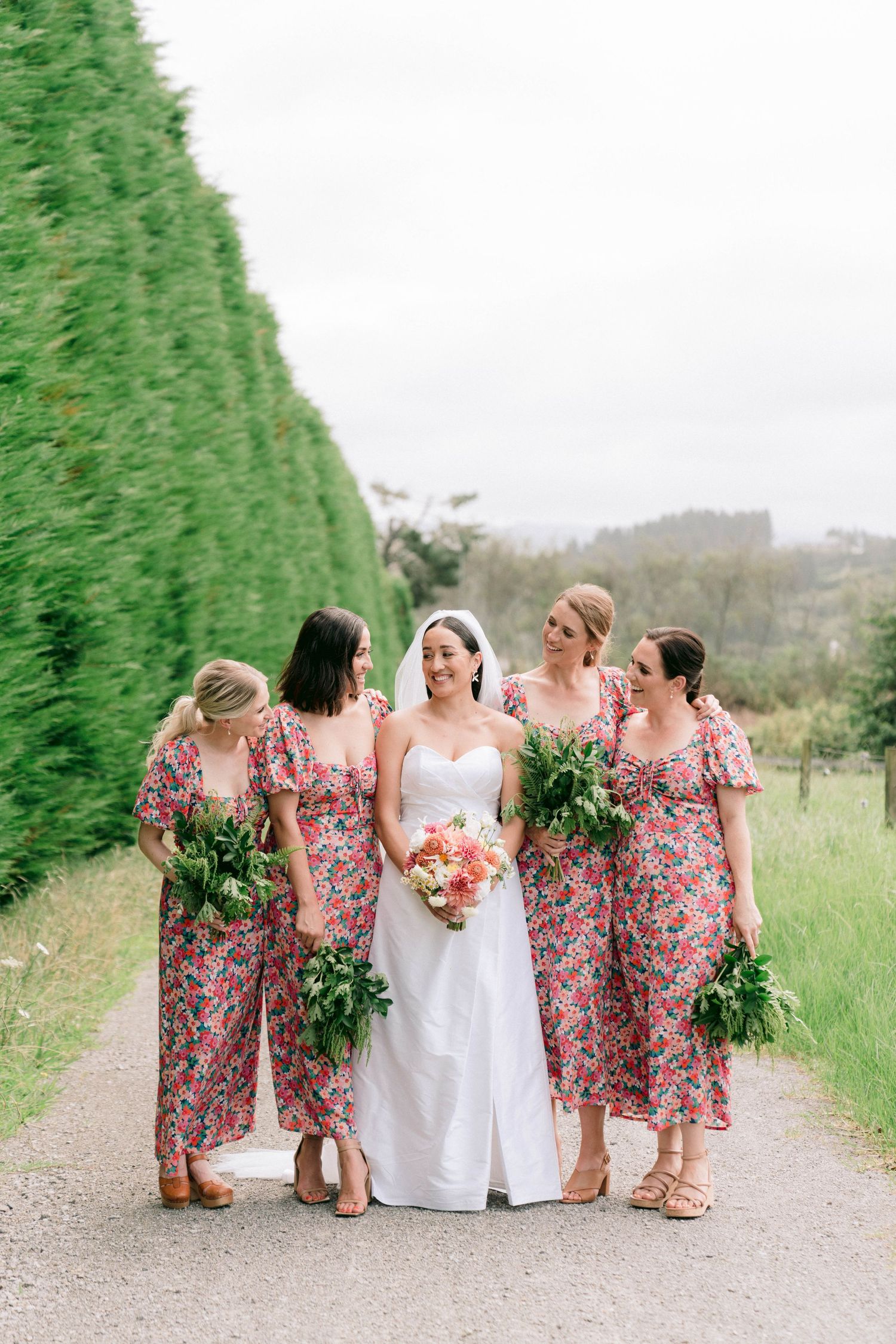 Bridesmaids in floral dresses carrying bouquets walk with bride along a garden path beside tall hedges.